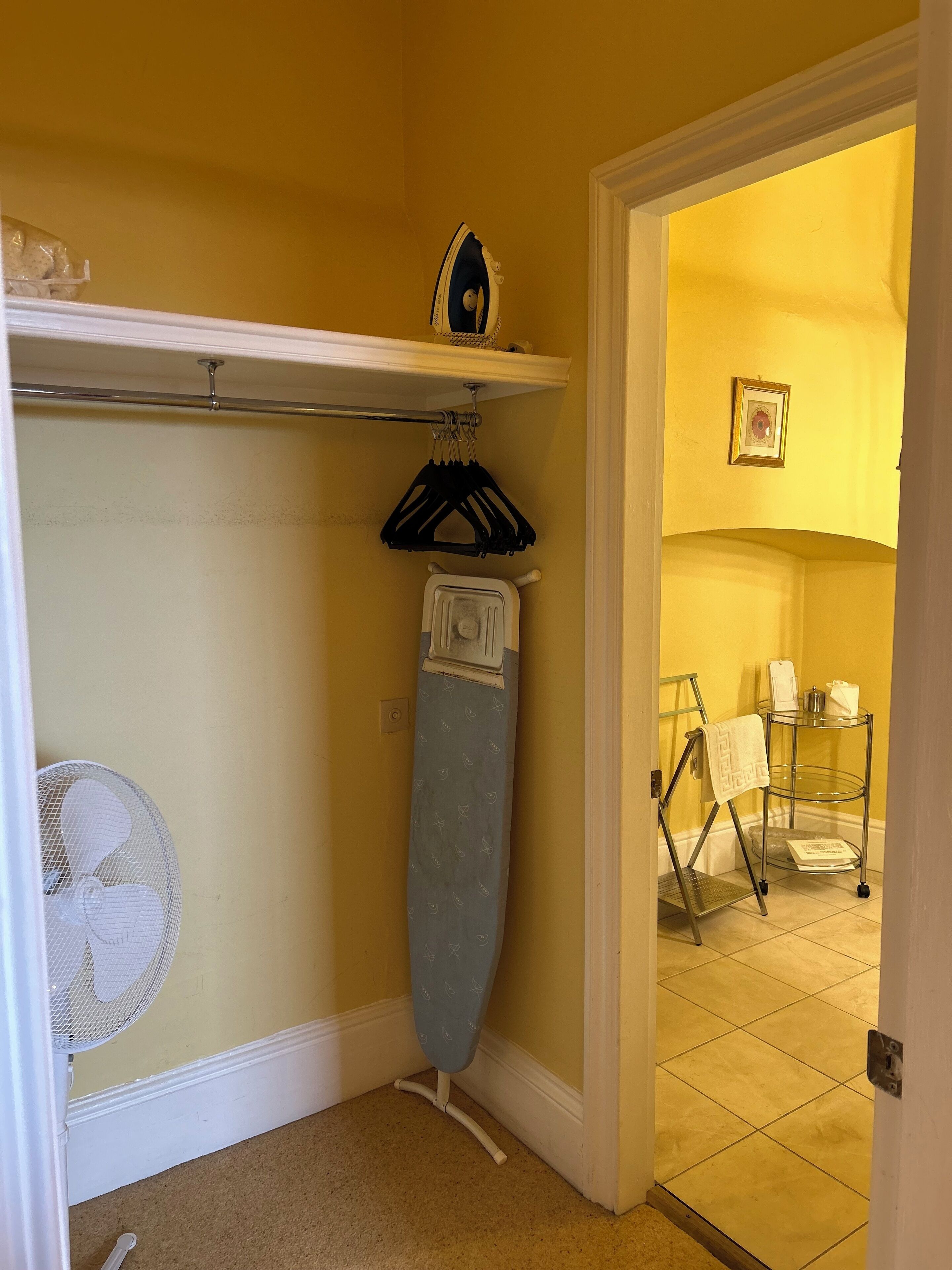 Un área de lavandería con una tabla de planchar, un ventilador y una repisa metálica con una toalla.