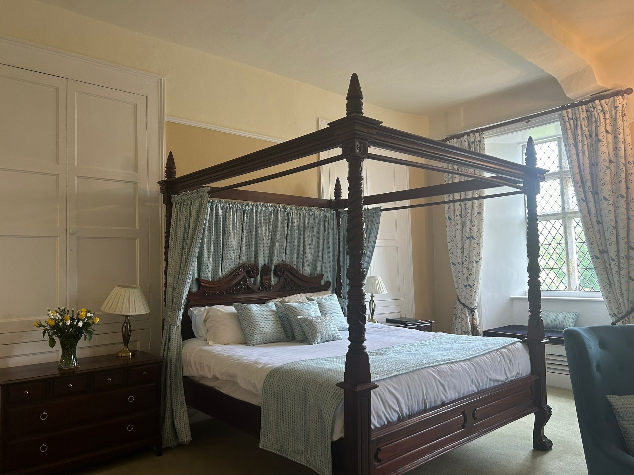 A four-poster canopy bed with a wooden frame, a dresser with a vase of flowers, and a window with curtains.