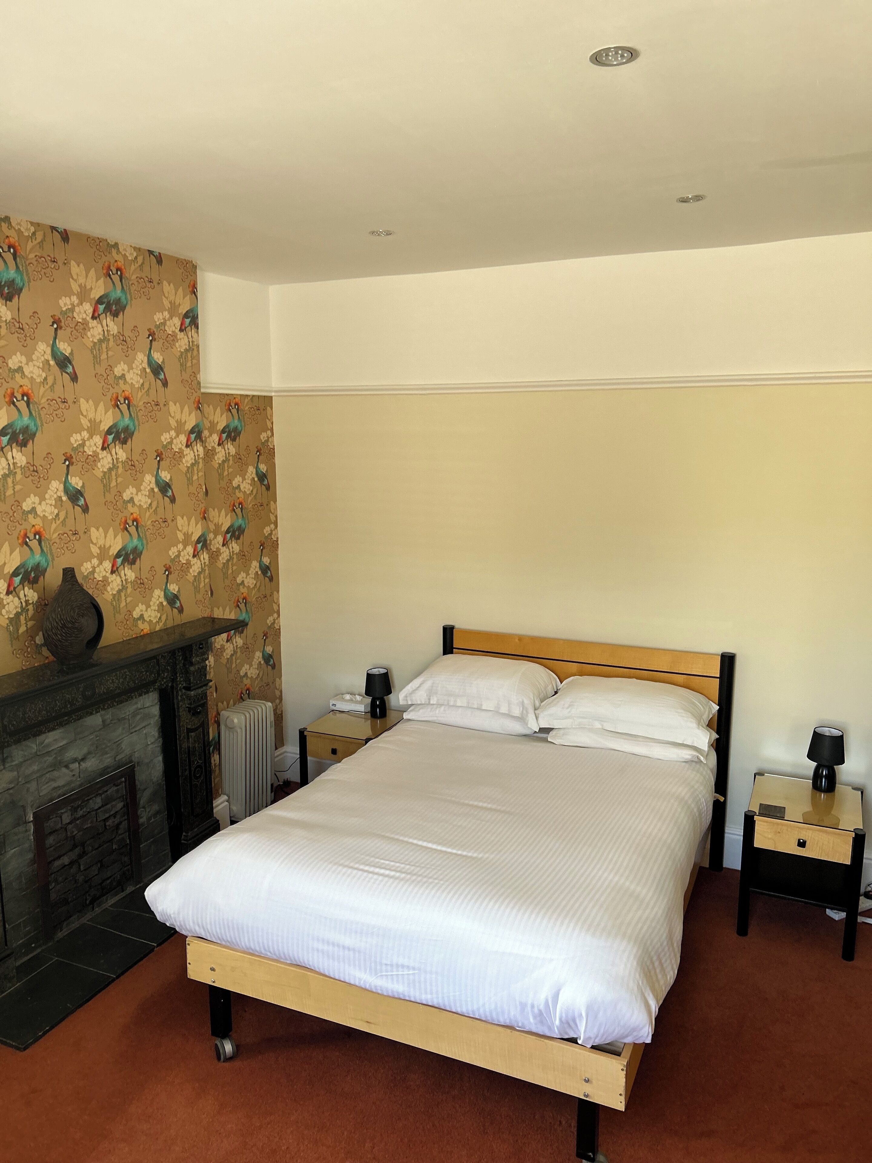 A neatly made bed with white linens, a wooden headboard, and two bedside tables with lamps. A decorative vase is on the mantel above a fireplace.