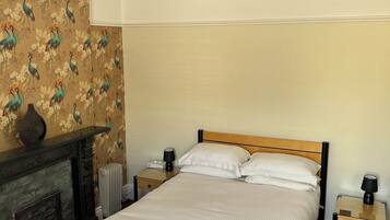 A neatly made bed with white linens, a wooden headboard, and two bedside tables with lamps. A decorative vase is on the mantel above a fireplace.