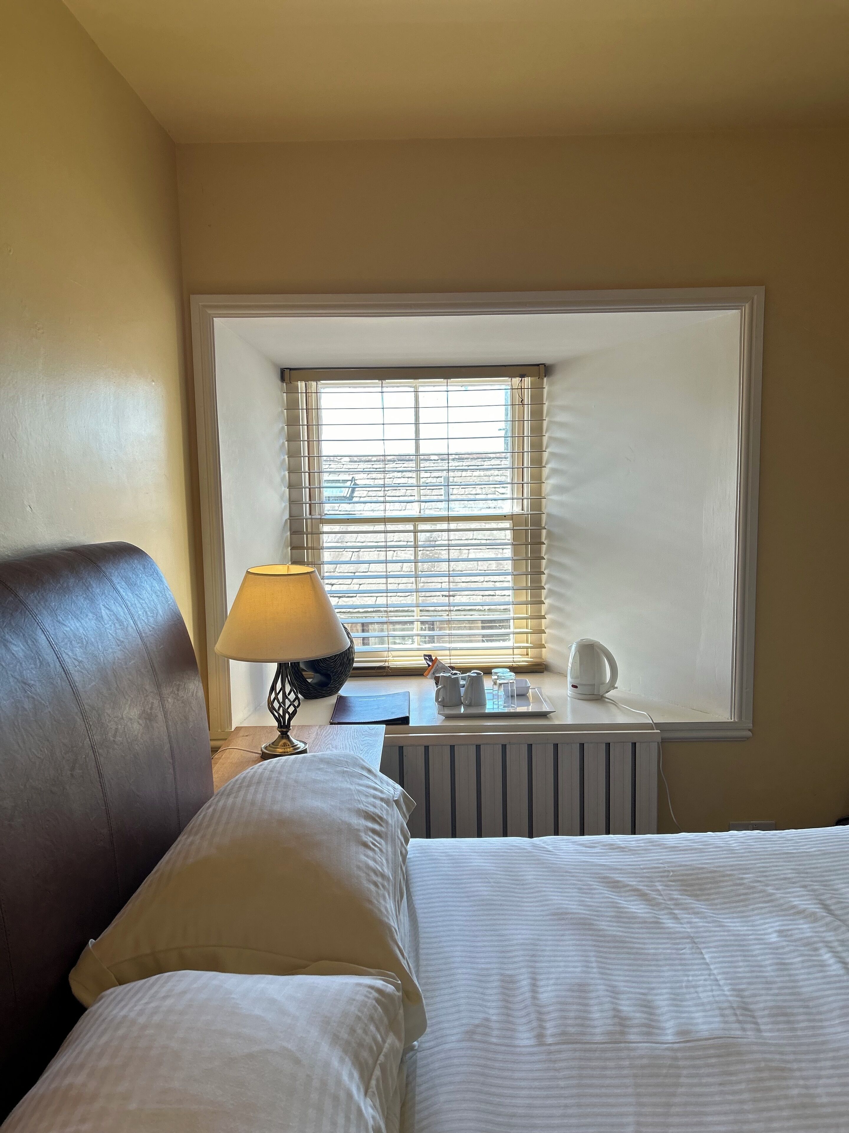 A hotel room with a bed, a bedside table with a lamp, a radiator, and a window with blinds.