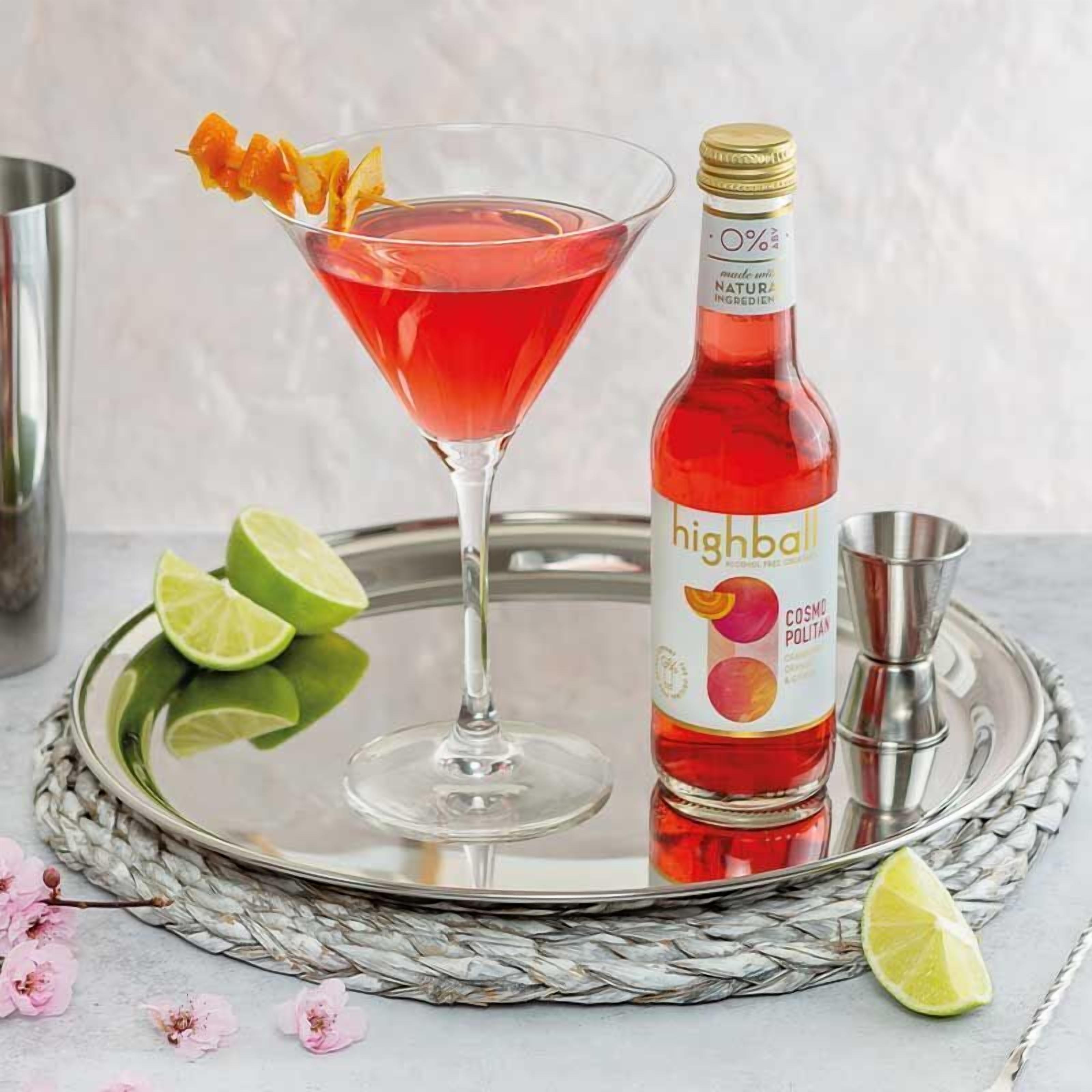 Una copa de martini con un cóctel rojo, una botella de highball y gajos de lima sobre una bandeja de plata.