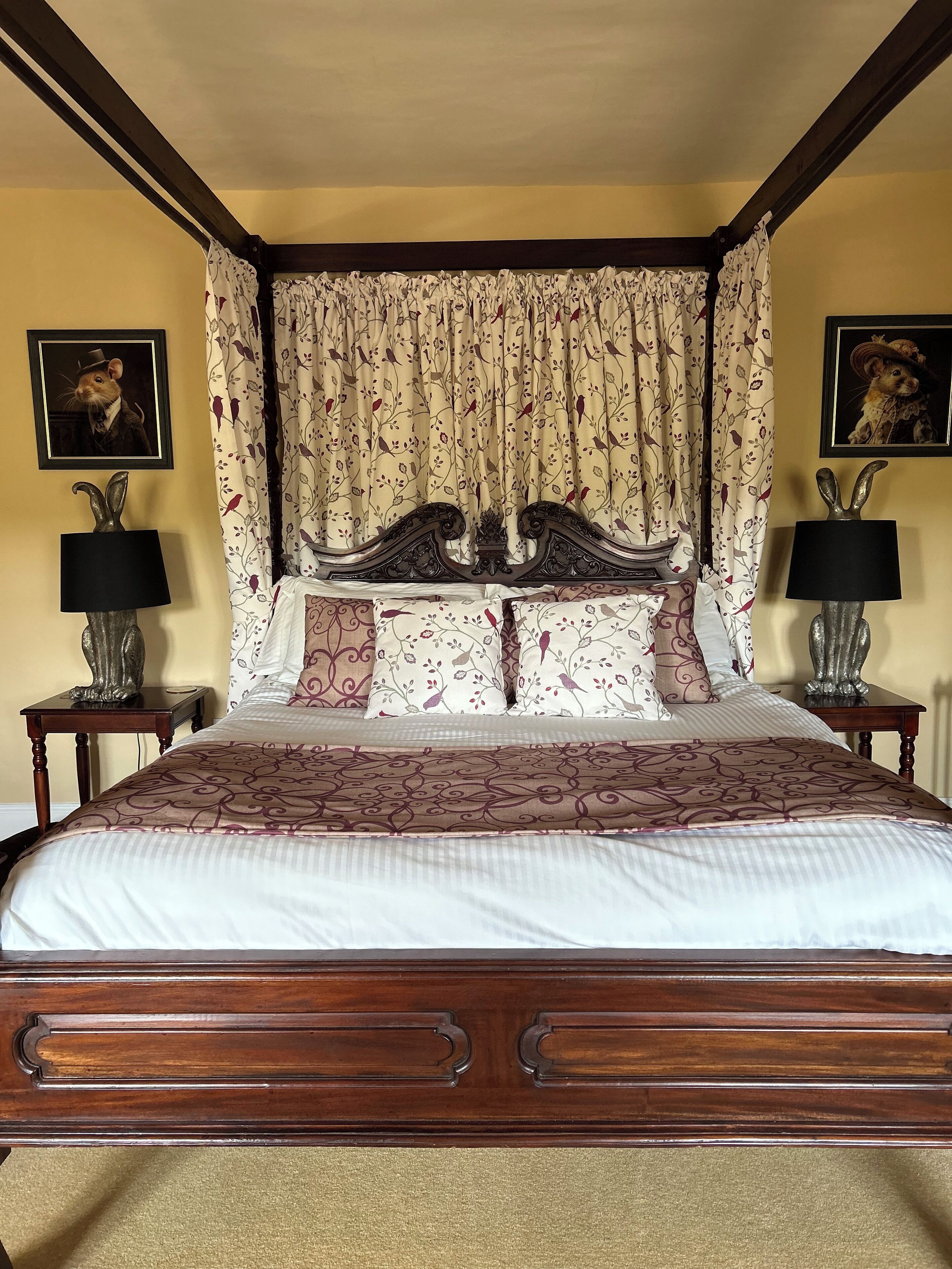 A four-poster bed with floral curtains, two nightstands with lamps, and two framed pictures on the walls.