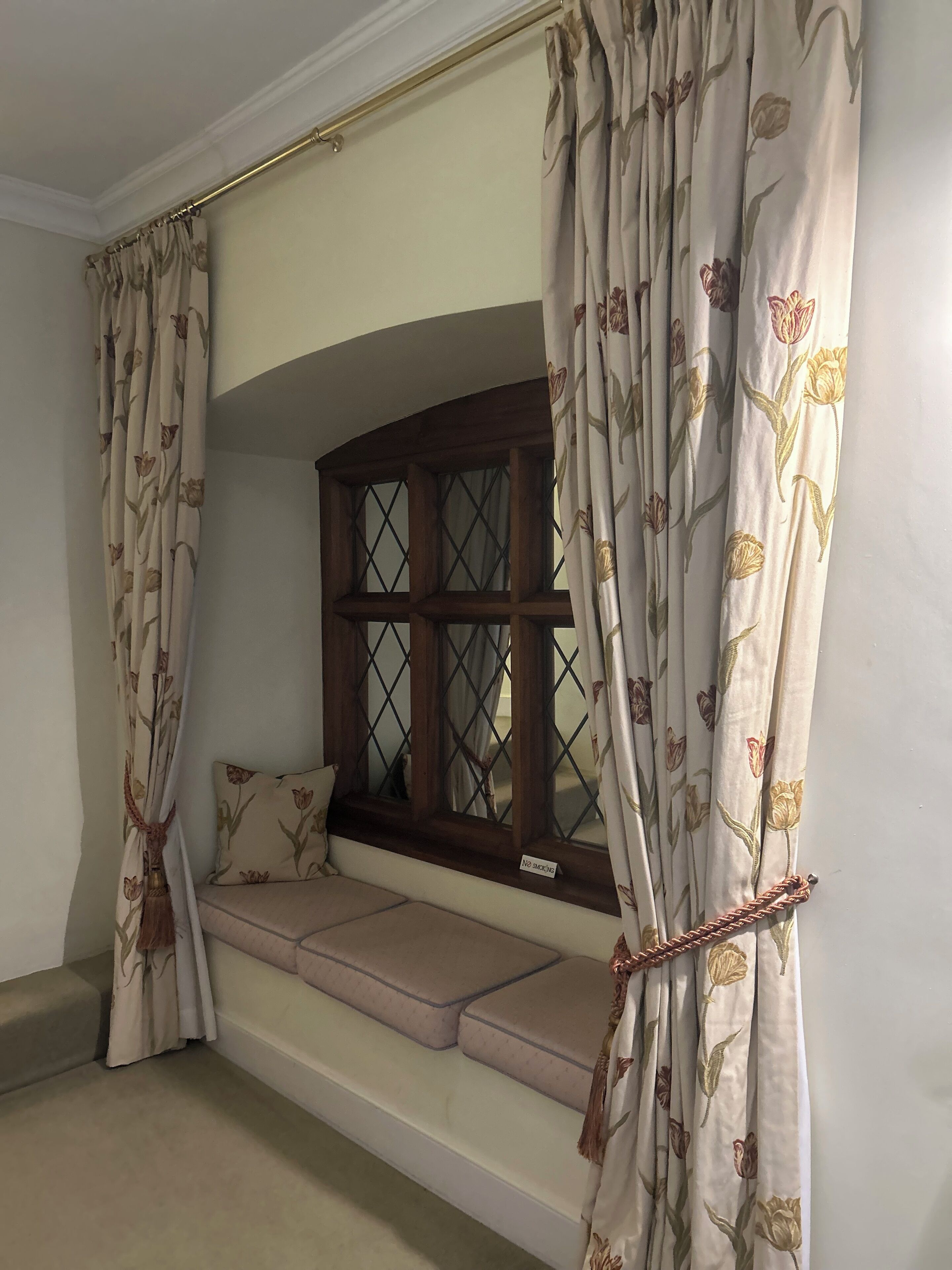 Una habitación con asiento junto a la ventana, cortinas con motivos florales y un cartel de 'No Fumar'.