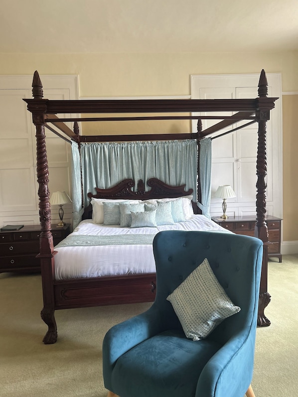 A four-poster bed with a canopy, a blue upholstered armchair, and a bedside table with a lamp.