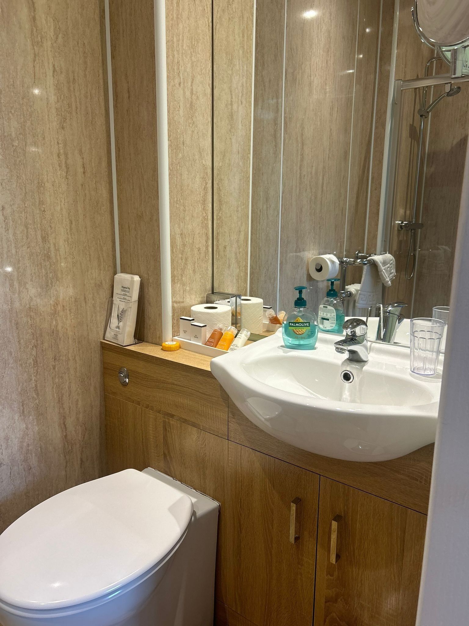 Et moderne badeværelse med hvid vask, spejl og toilet. Der er toiletartikler på bordet, og et håndklæde hænger på brusekabinedøren.