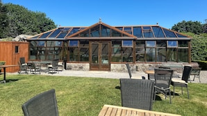 A large glass conservatory with a wooden frame, a stone foundation, and a wooden deck with outdoor furniture. - Moresby Hall (Whitehaven)