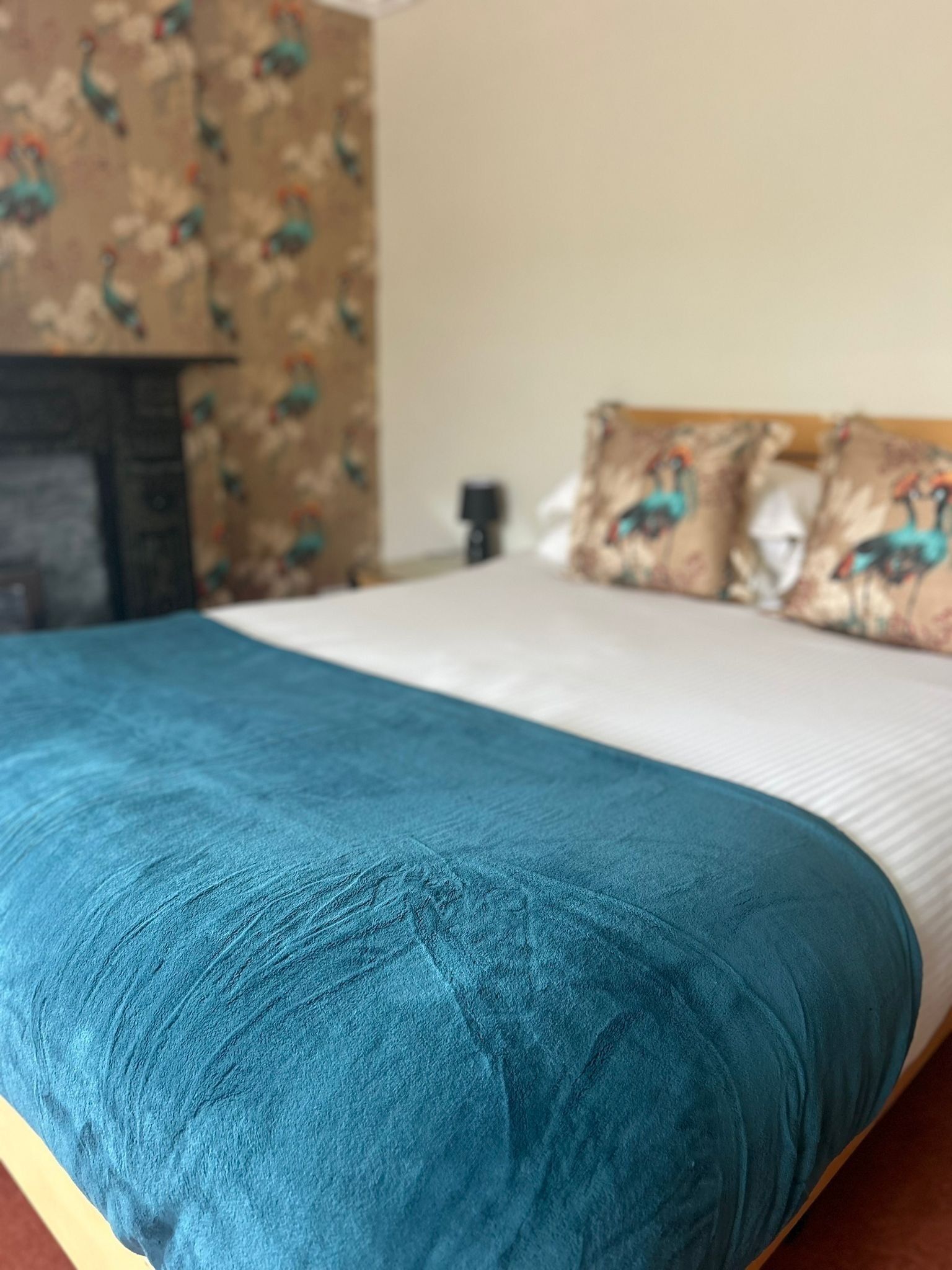 Una cama con colcha color aguamarina y almohadas blancas, una mesita de noche con lámpara y una pared con estampado floral.