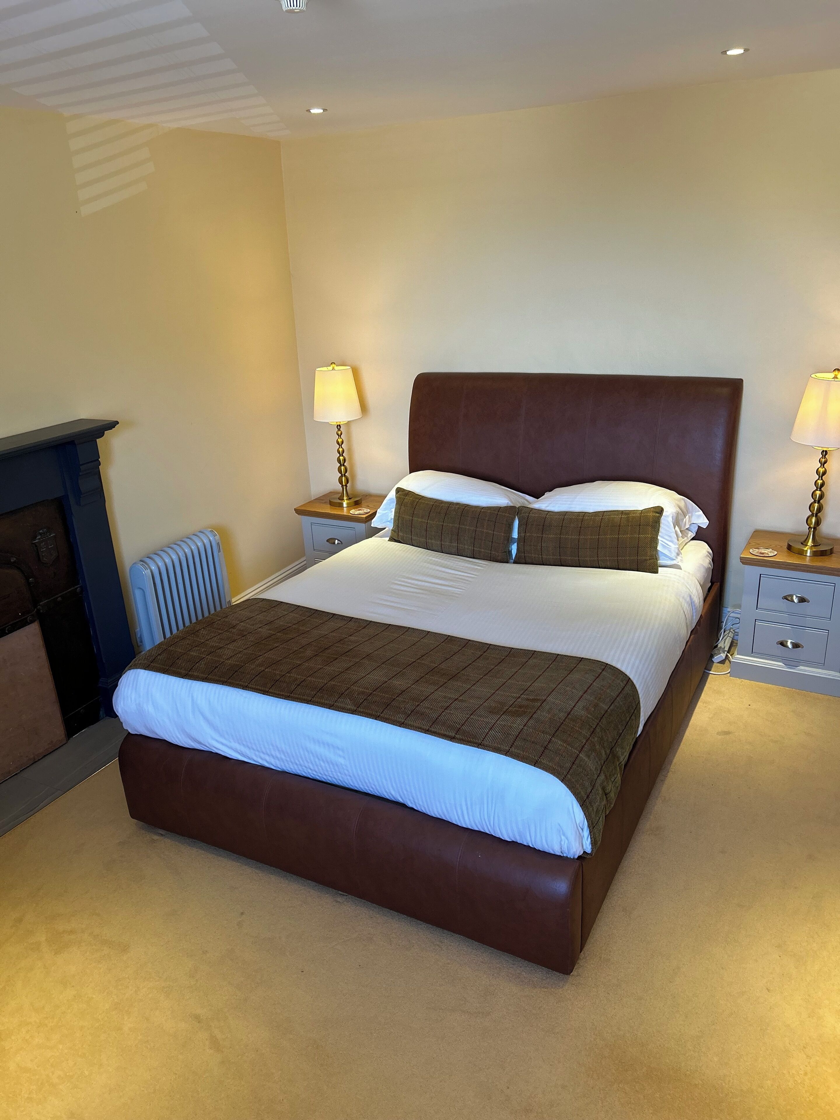 A neatly made bed with a brown headboard, two bedside tables with lamps, a radiator, and a fireplace.