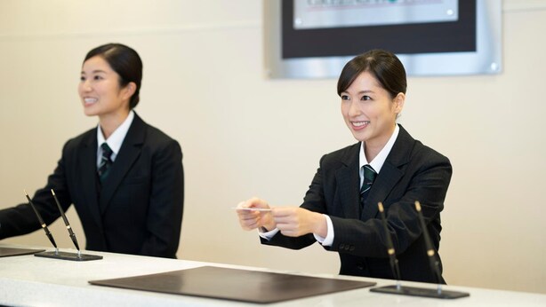 Two individuals in business attire at a conference table with microphones, in a room with a sign reading "GREENSHOTELS". - Hotel Econo Tsu Station (Tsu)