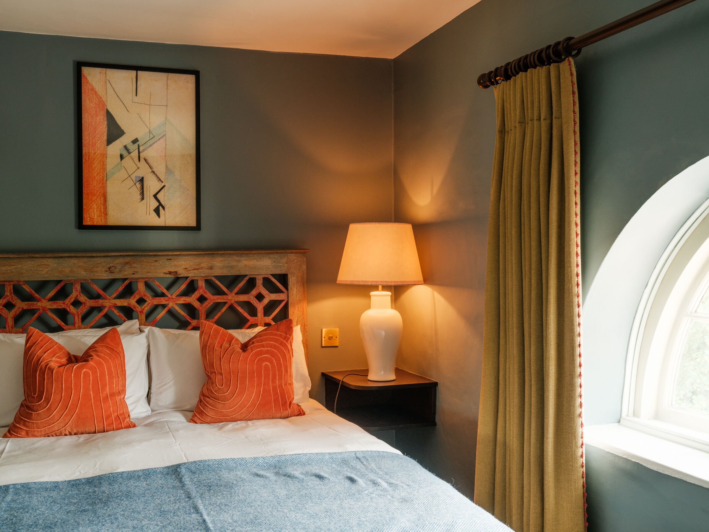 A hotel room with a bed, a nightstand, a lamp, a window, and a painting on the wall.