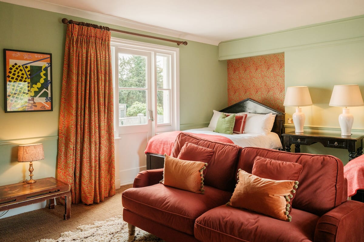 A room with a bed, a red sofa, a small table with a lamp, and a window with curtains.