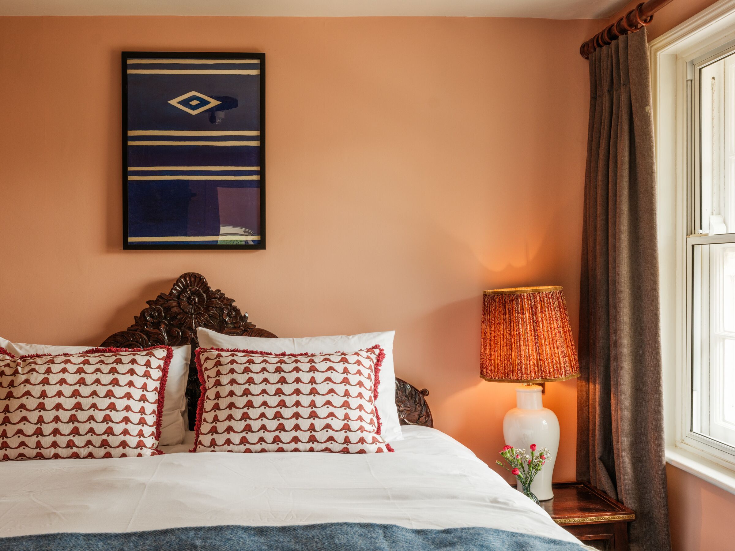 A neatly made bed with patterned pillows, a decorative headboard, a framed artwork on the wall, a bedside lamp, and a window with curtains.