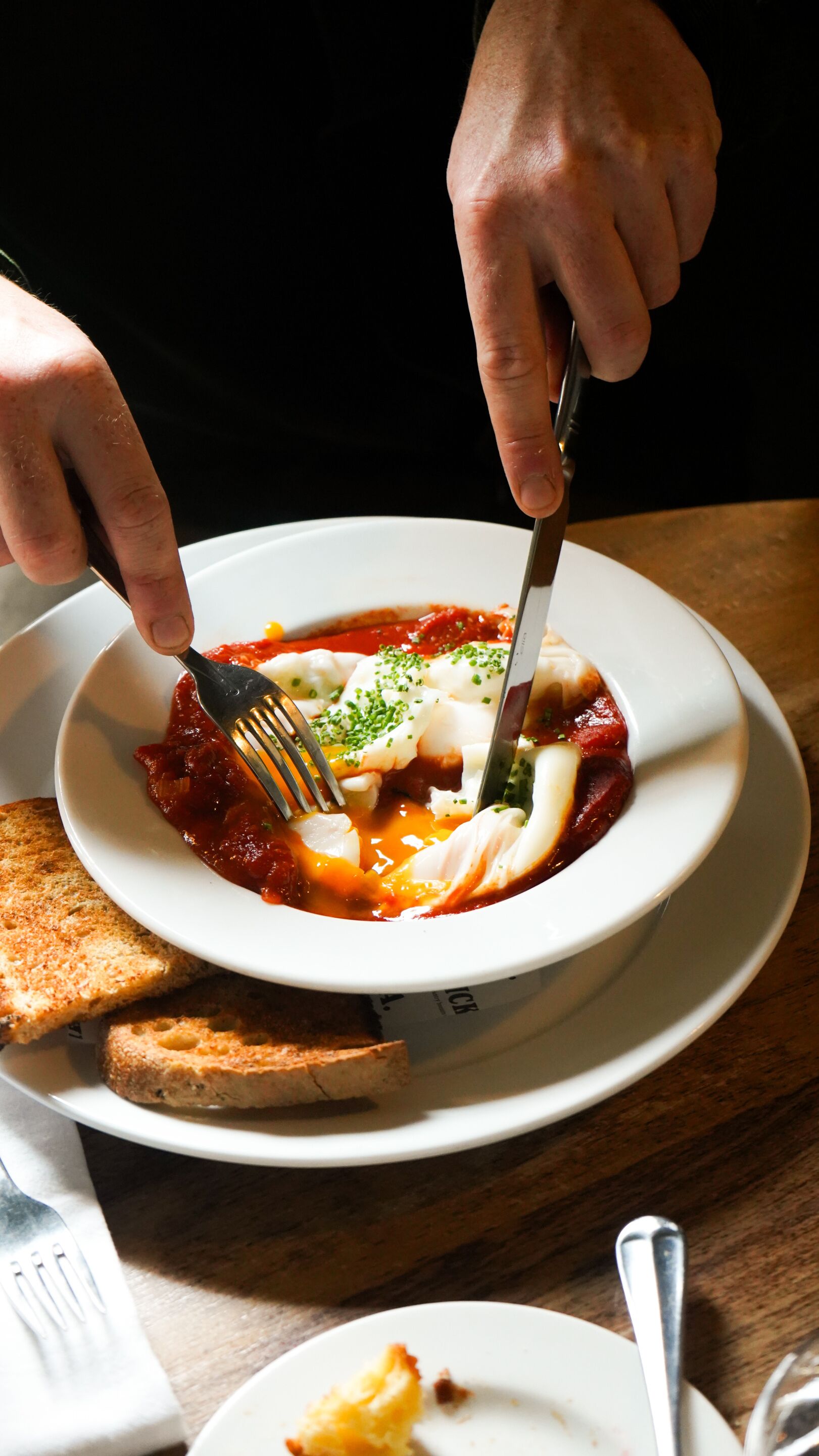 A person eating a dish with a fork and knife, with toast on the side.