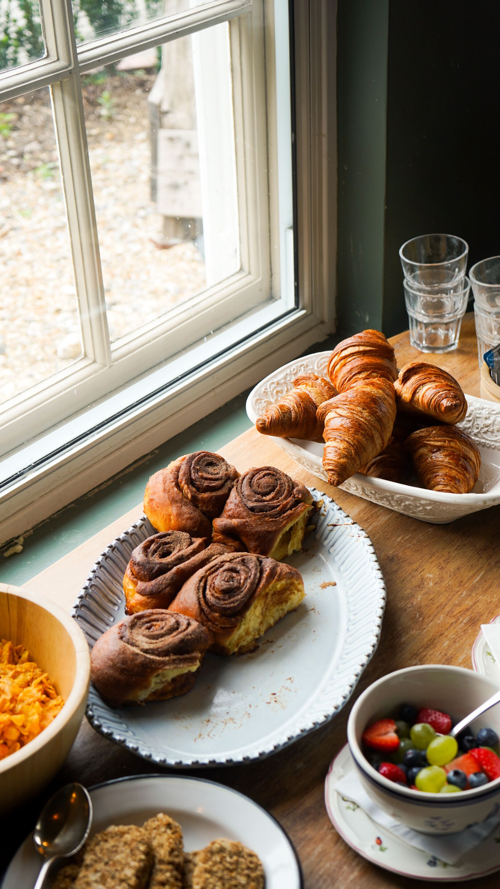 Une table de petit-déjeuner avec des croissants, des pains aux cannelle et un saladier de fruits.