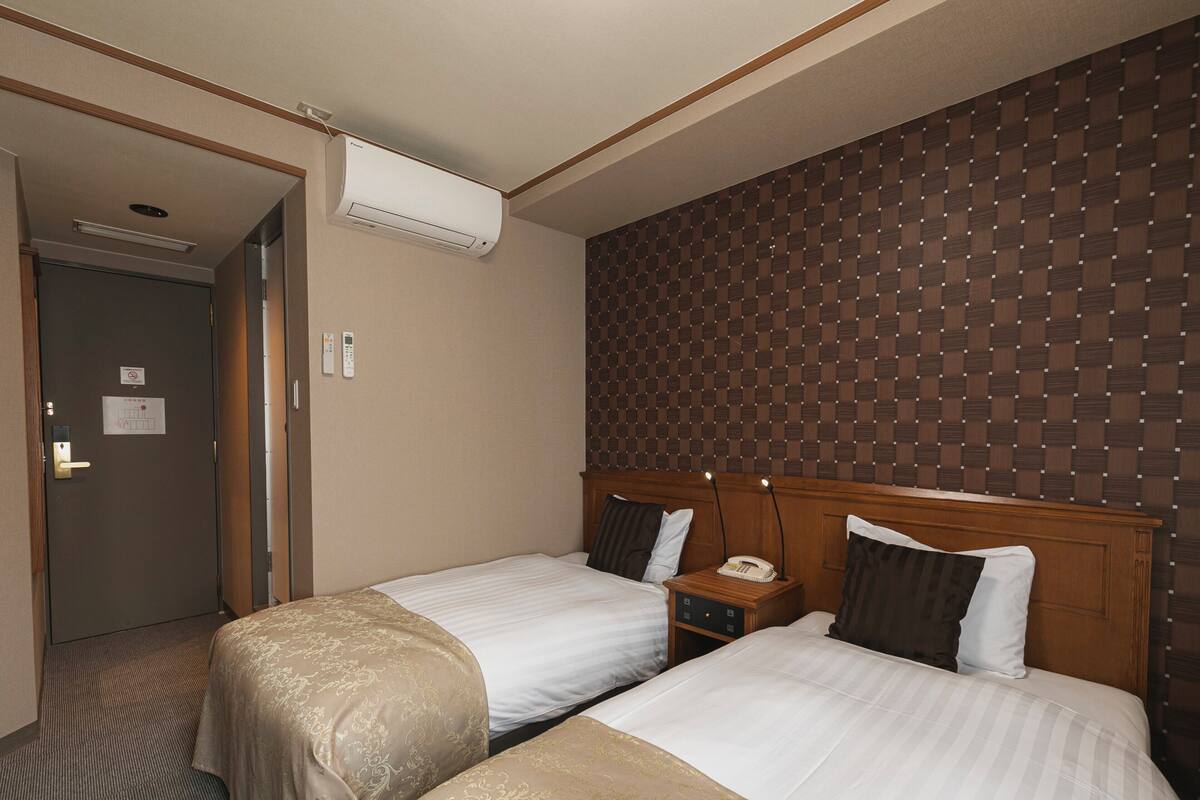 A hotel room with two beds, a wooden headboard, a bedside table with a telephone, and an air conditioning unit.
