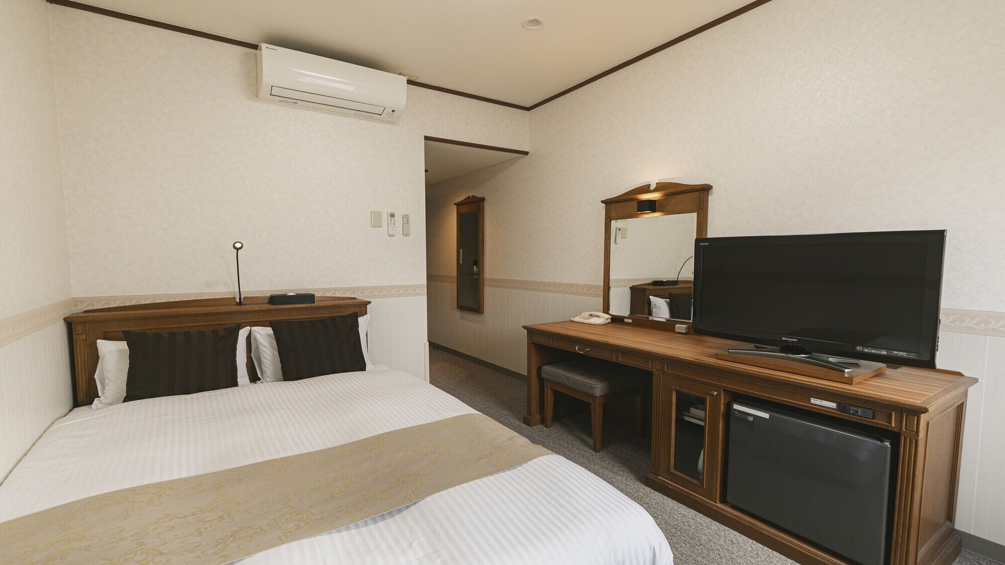 A hotel room with a bed, a desk with a television, a minibar, and an air conditioning unit.