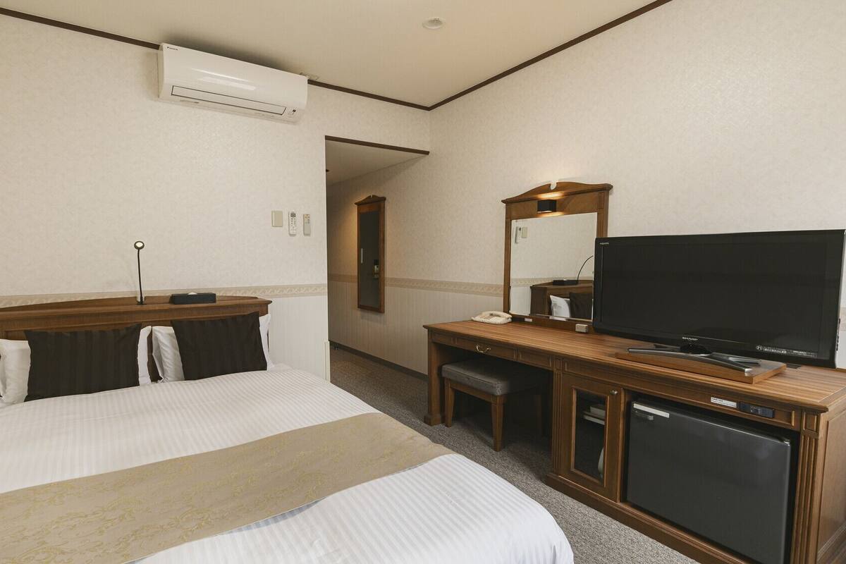 A hotel room with a bed, a desk with a television, a minibar, and an air conditioning unit.