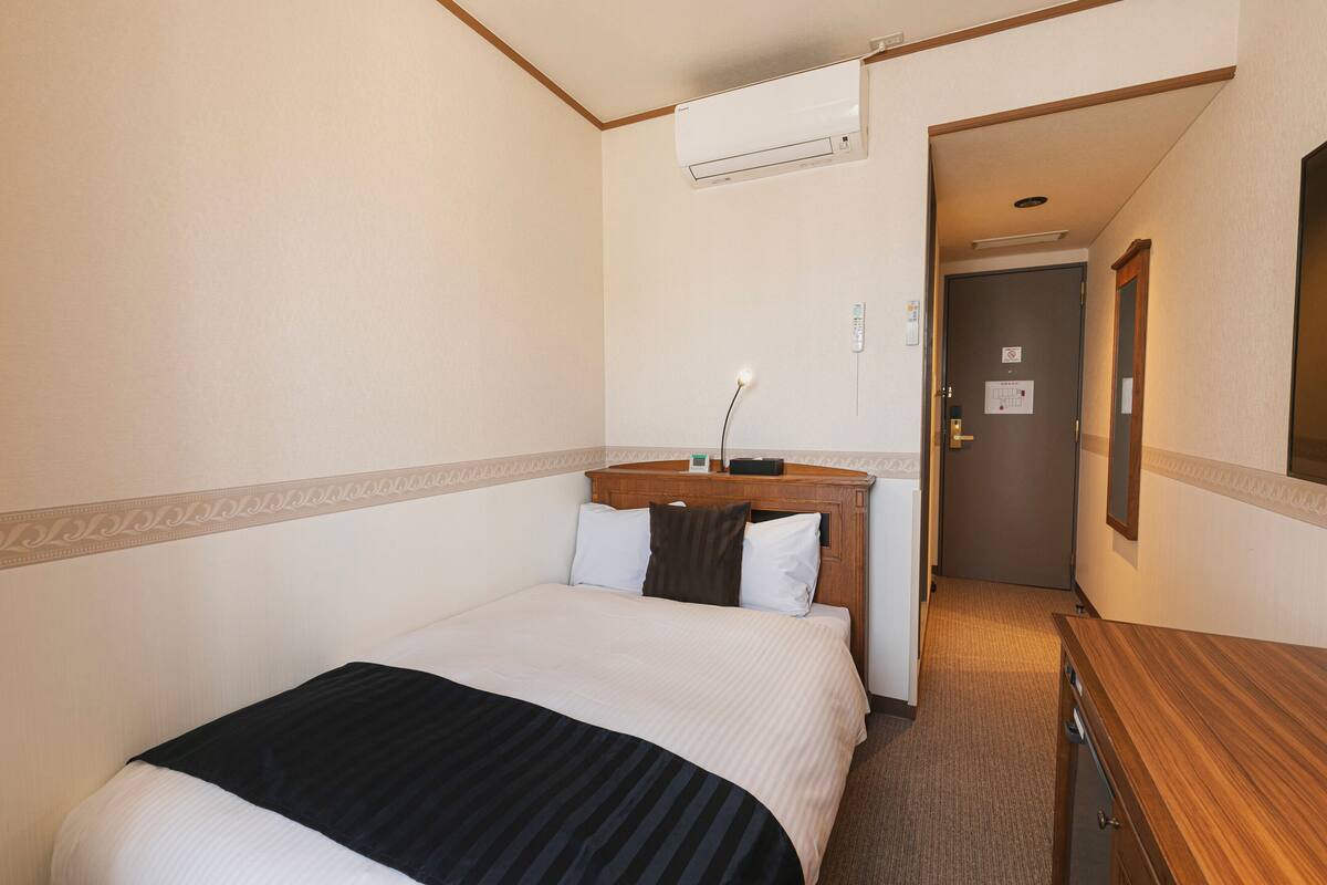 A hotel room with a bed, bedside table, lamp, and an air conditioning unit.