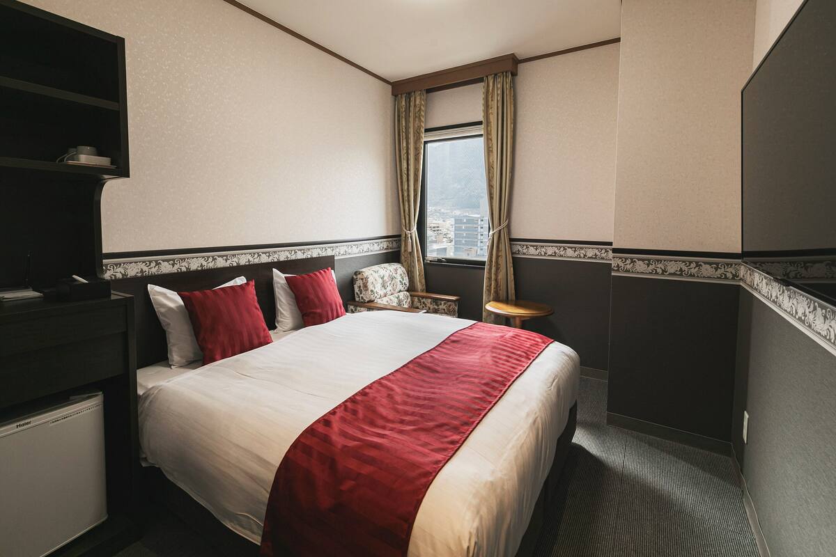 A hotel room with a bed, a desk, a chair, a window with curtains, and a view of the city.