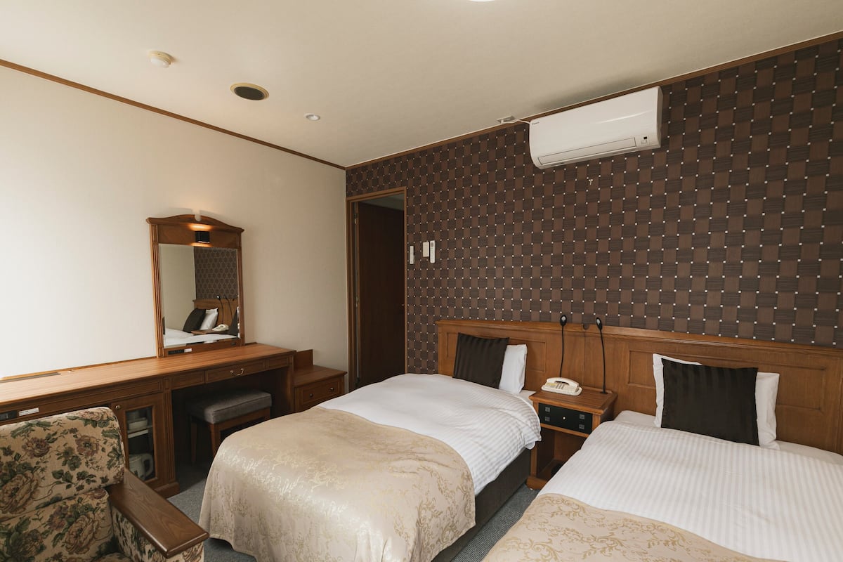 A hotel room with two beds, a desk with a mirror, a chair, and an air conditioning unit on the wall.