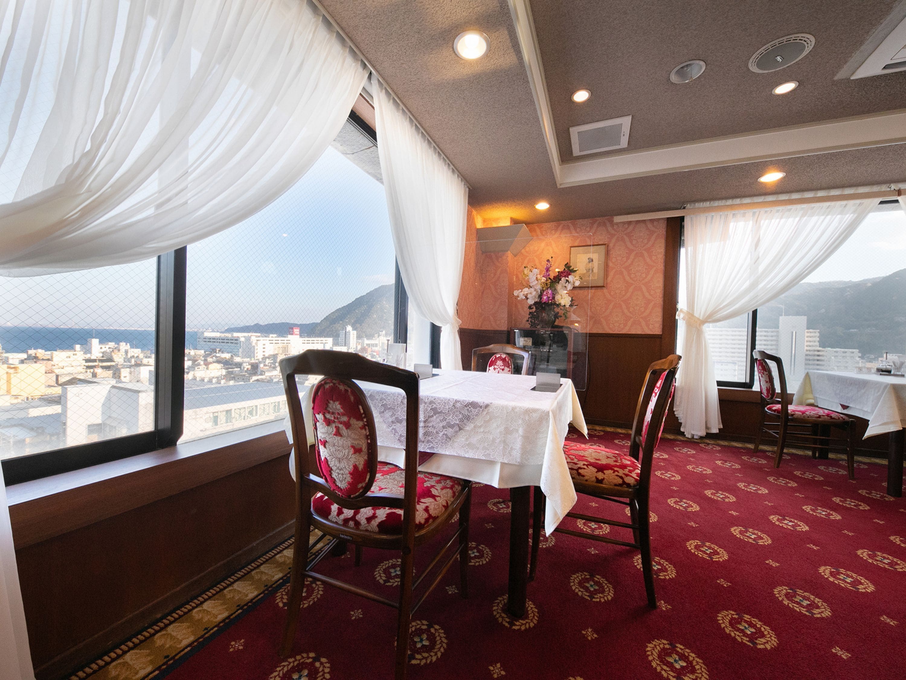 A dining area with a table set for two, chairs with floral upholstery, a red patterned carpet, and a city view through large windows with white curtains.