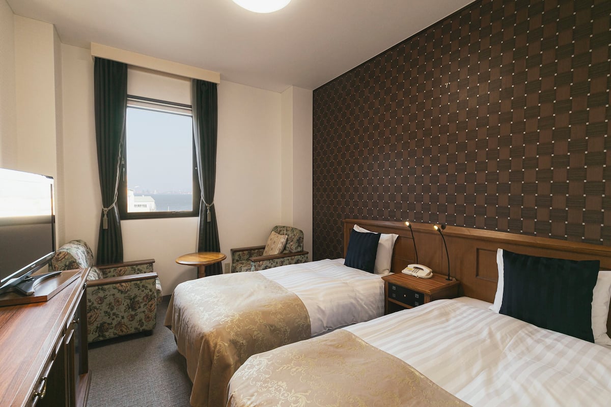A hotel room with two beds, a desk, a chair, a small table, and a window with curtains.