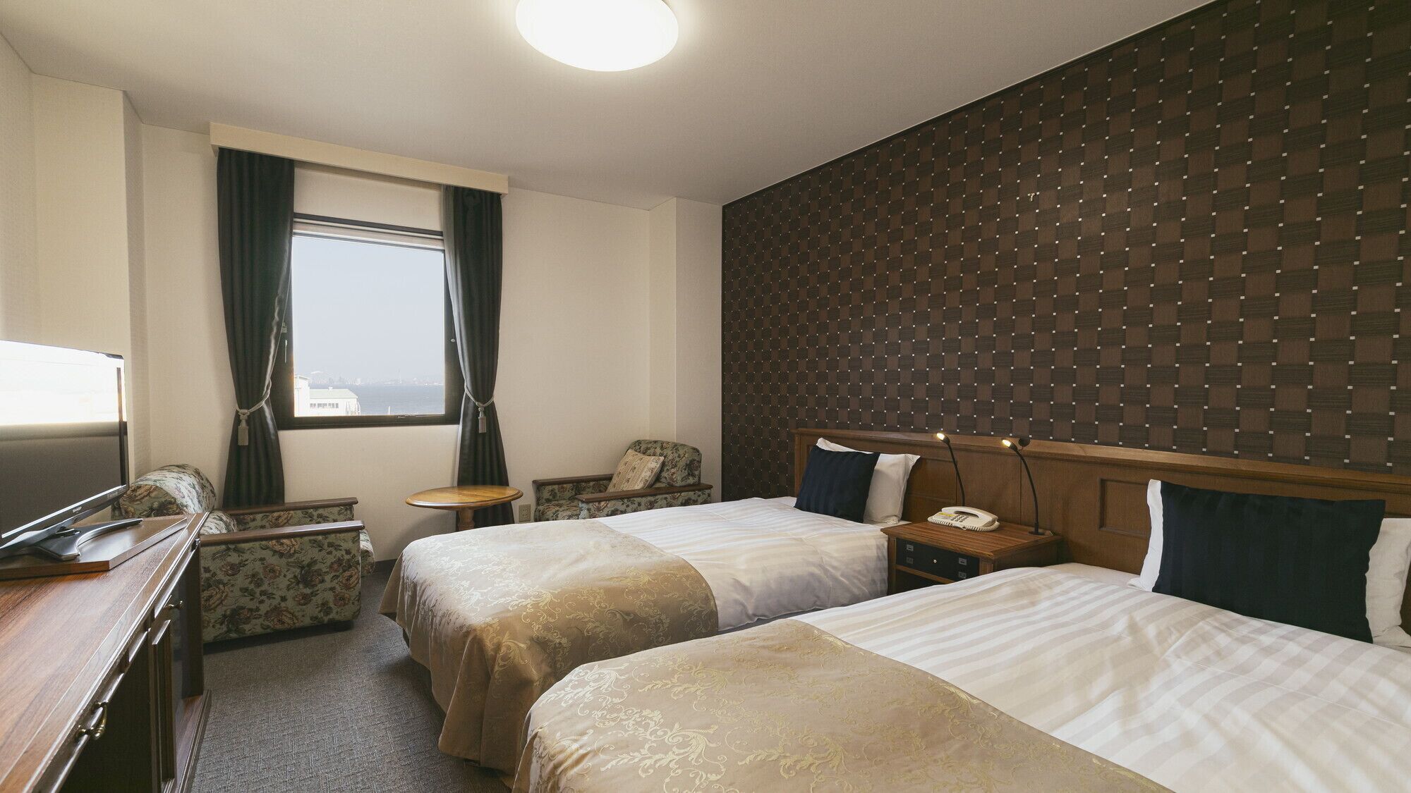 A hotel room with two beds, a desk, a chair, a small table, a telephone, and a window with curtains.