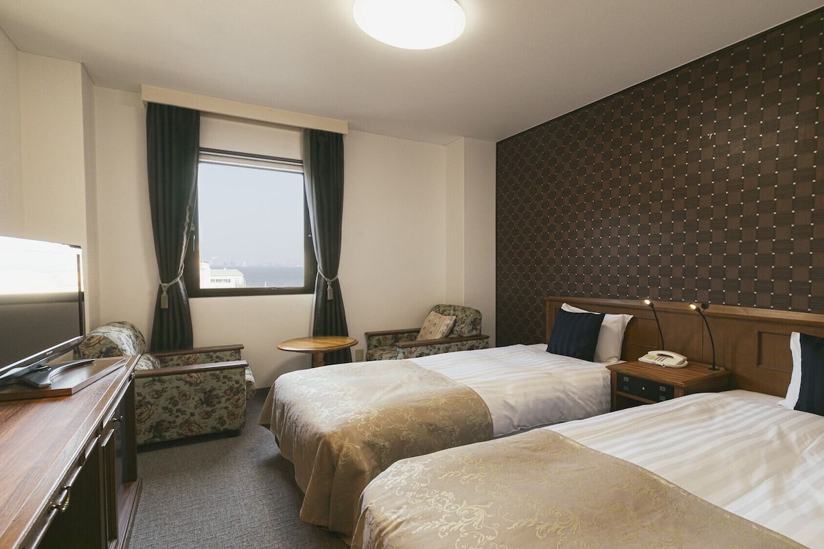 A hotel room with two beds, a desk, a chair, a small table, a telephone, and a window with curtains.