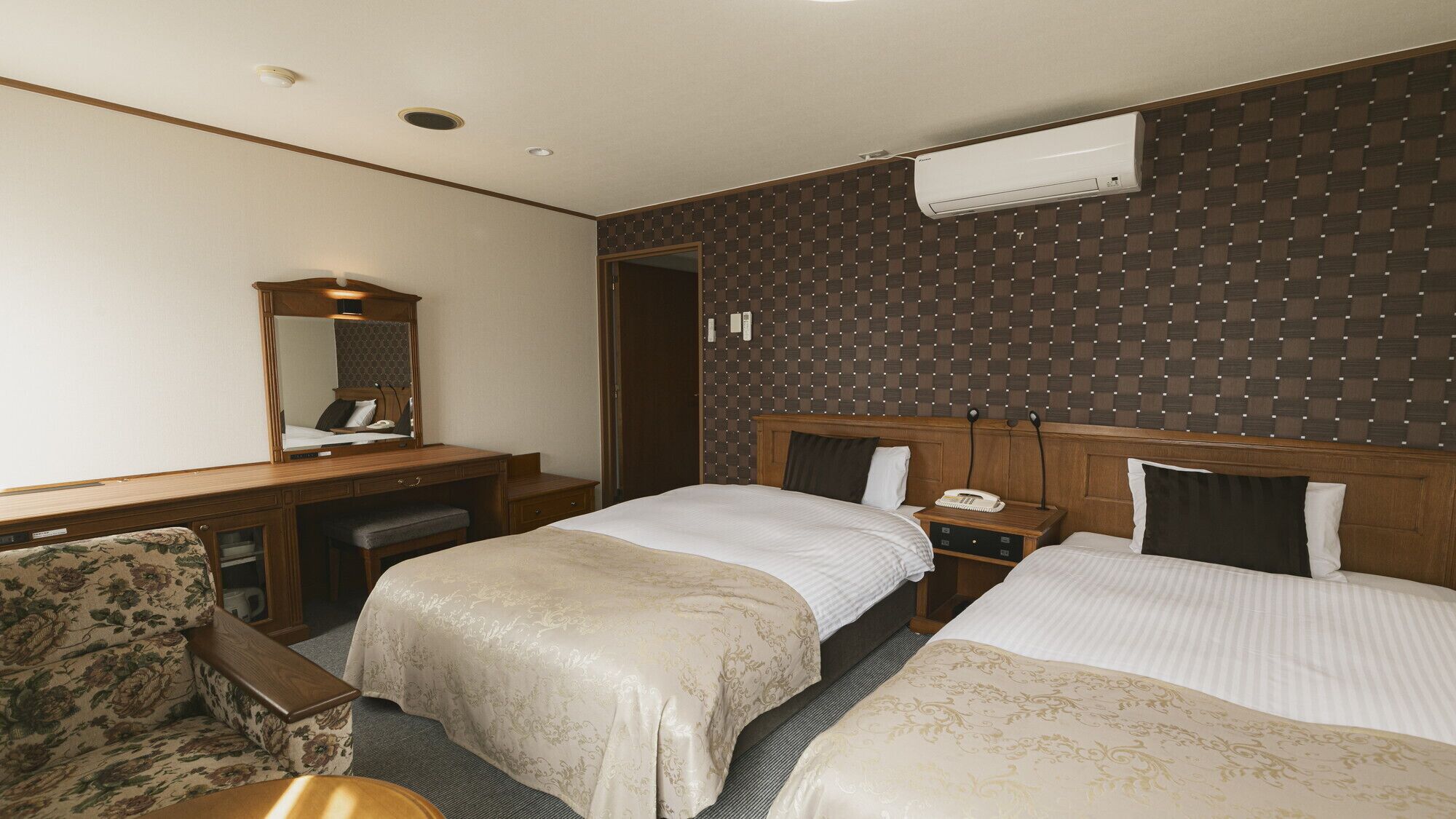 A hotel room with two beds, a desk, a chair, a mirror, and an air conditioning unit.