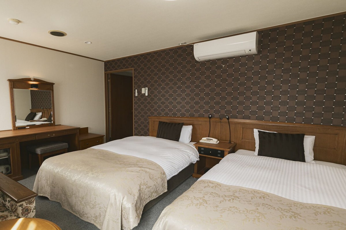 A hotel room with two beds, a desk, a chair, a mirror, and an air conditioning unit.
