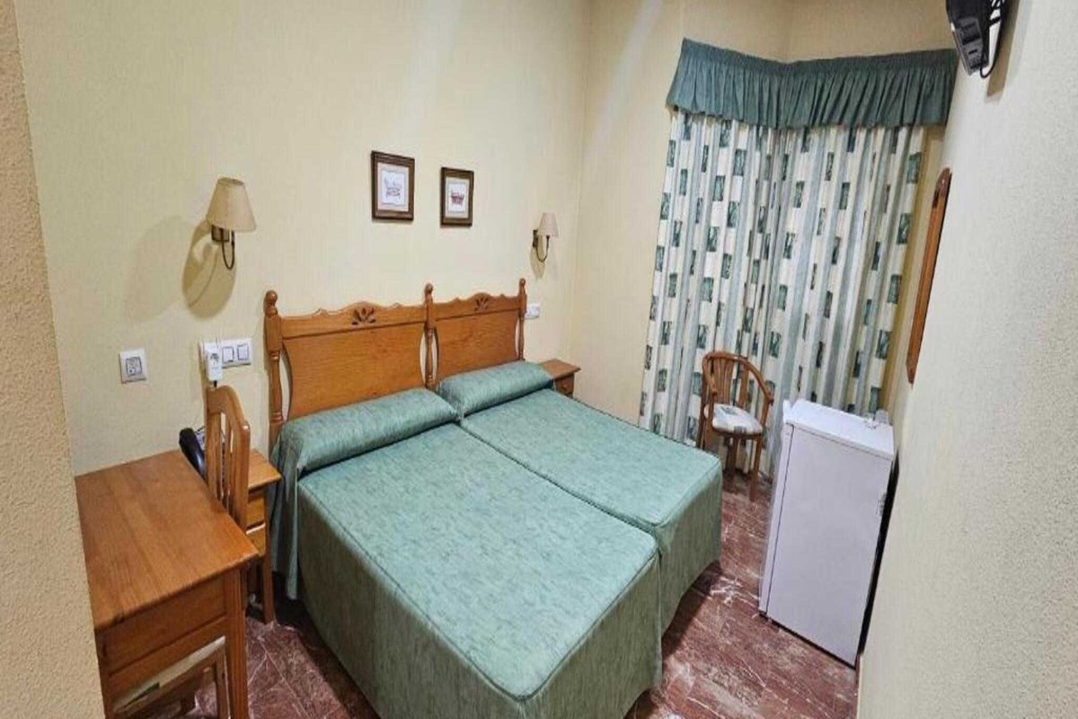 Cama doble con cabecera de madera, una mesita de noche con lámpara, una silla y un refrigerador.