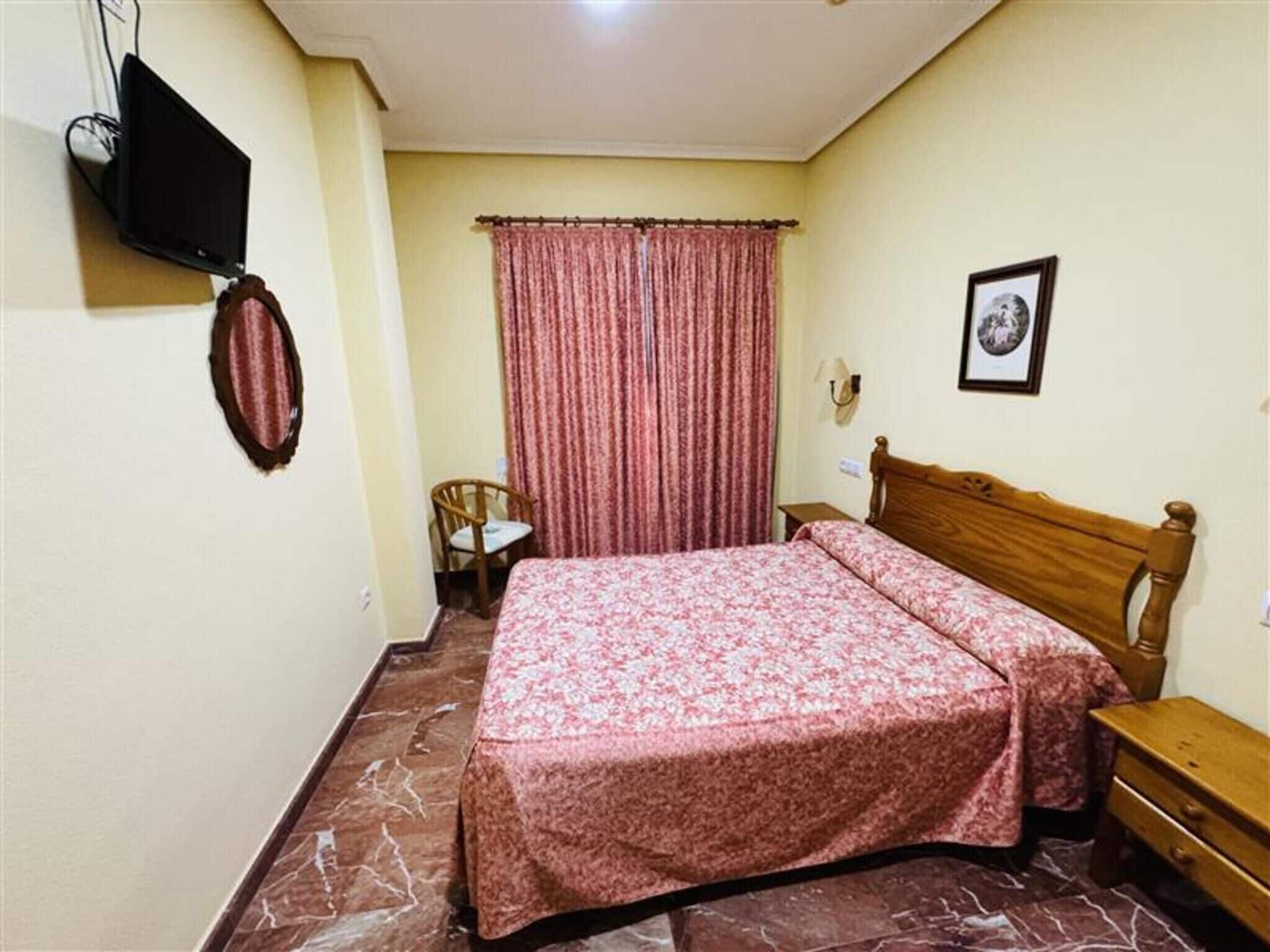 A bedroom with a bed, a chair, a small table, a television, and a framed picture on the wall.