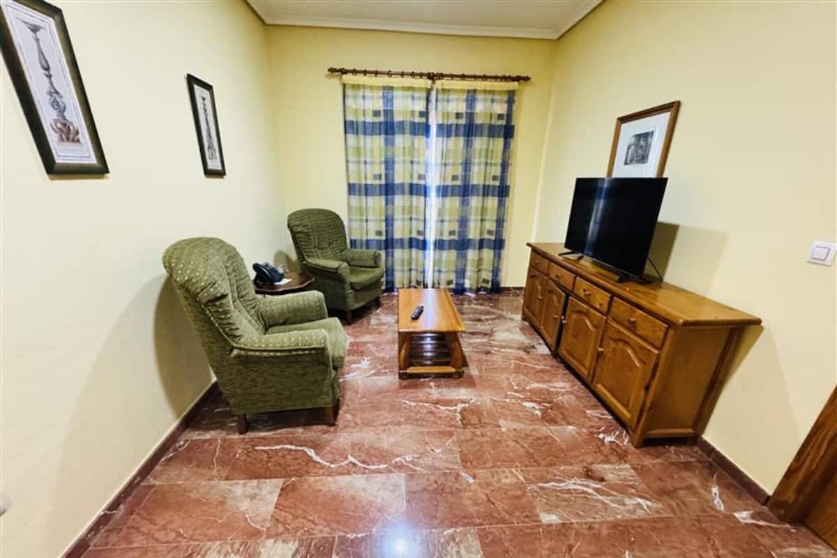Una sala de estar con dos sillones verdes, una mesa de centro de madera, un televisor sobre un mueble de madera y una cortina a cuadros.