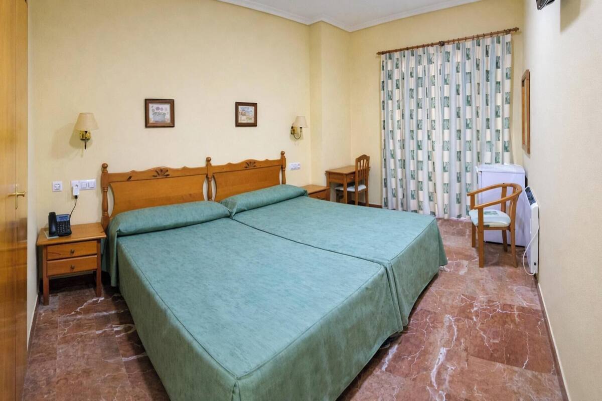 Una cama doble con cabecera de madera, dos mesitas de noche, una silla, un escritorio, una lavadora y una cortina.
