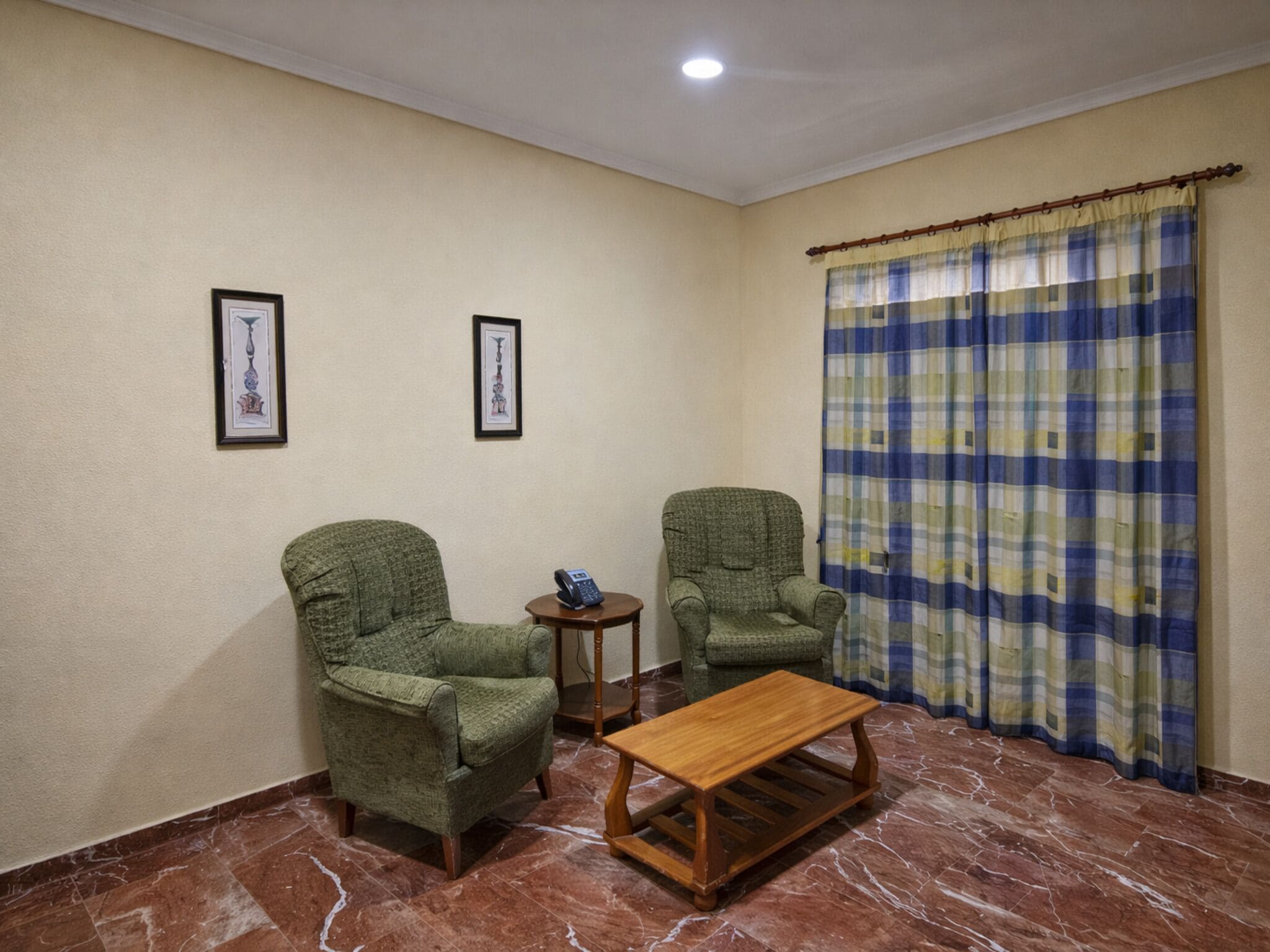 Une pièce avec deux fauteuils verts, une table basse en bois, une petite table d’appoint avec un téléphone et un rideau à carreaux.