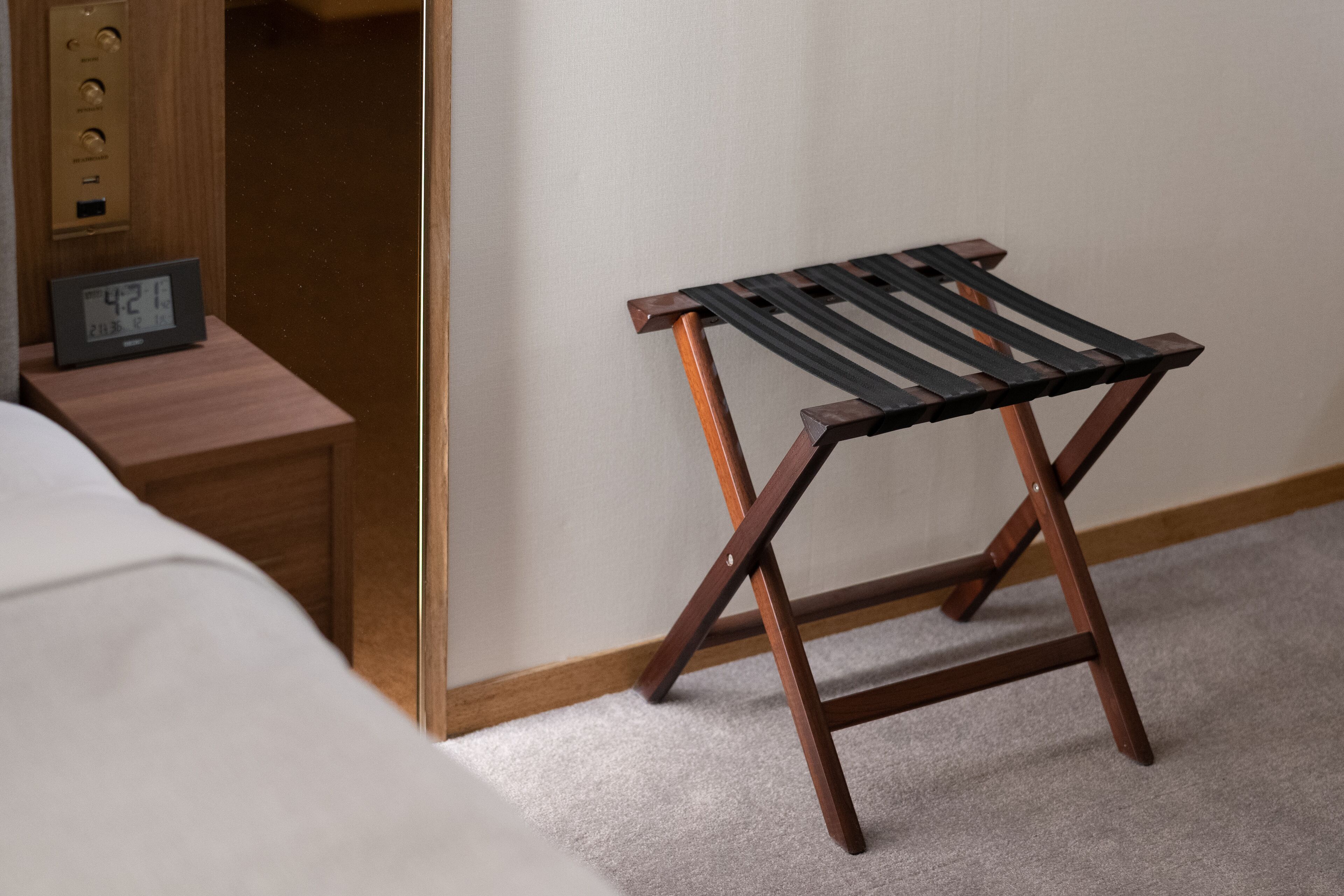 Un taburete de madera con asiento negro, una mesita de noche de madera con un reloj digital y una cama blanca en una habitación de hotel.