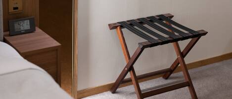 Un taburete de madera con asiento negro, una mesita de noche de madera con un reloj digital y una cama blanca en una habitación de hotel.