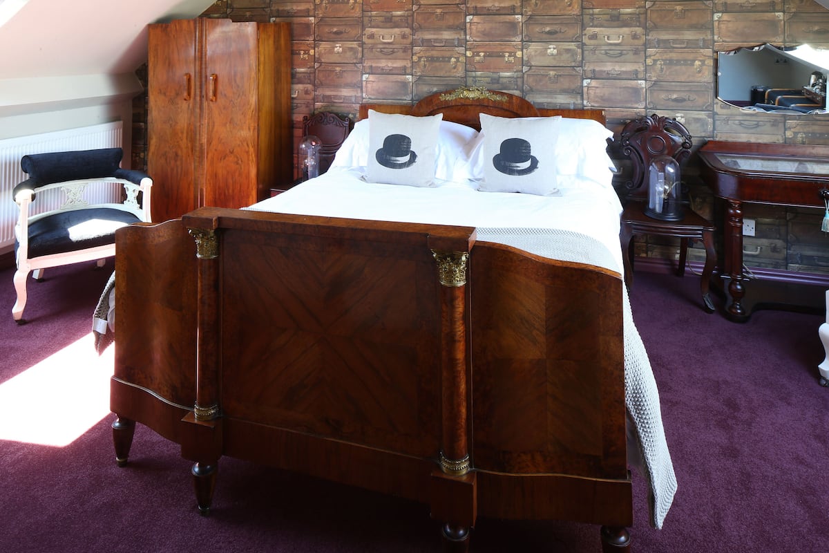 A bedroom with a wooden bed, a wardrobe, a mirror, and a wall decorated with vintage suitcases.
