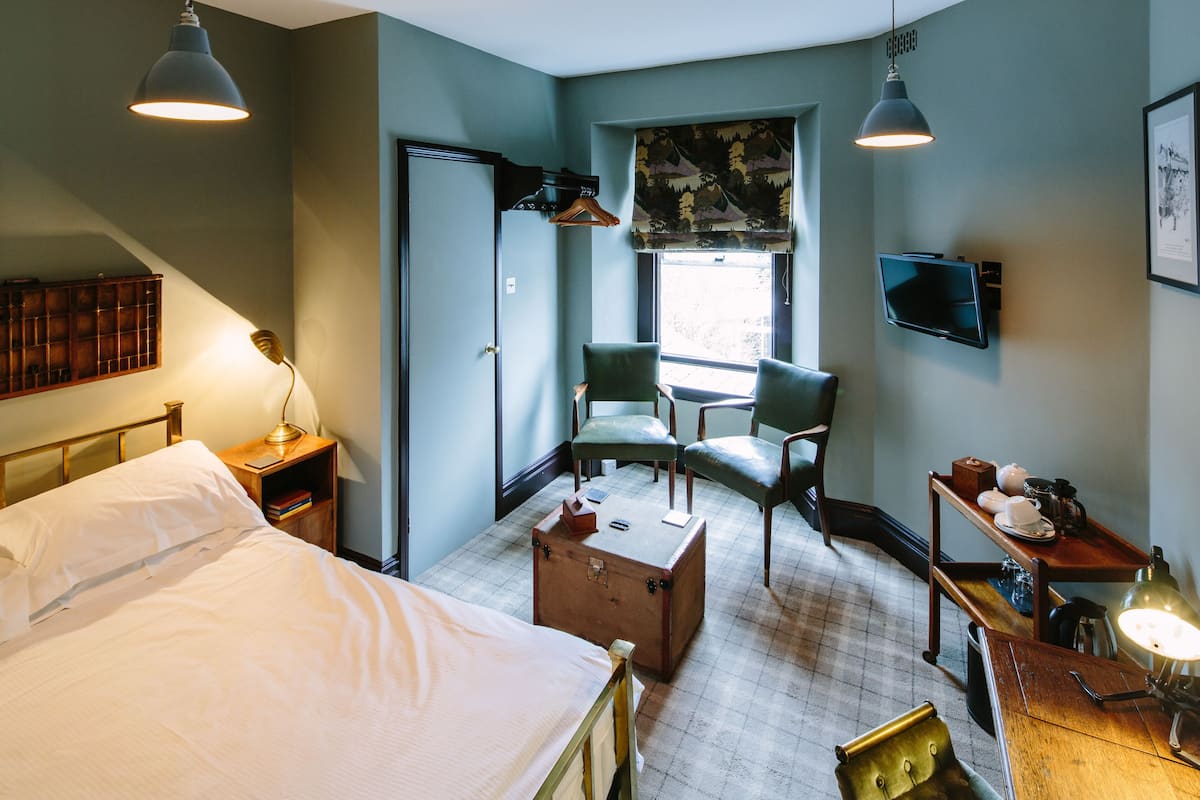 A bedroom with a bed, two chairs, a desk with a lamp, a suitcase, and a television on the wall.