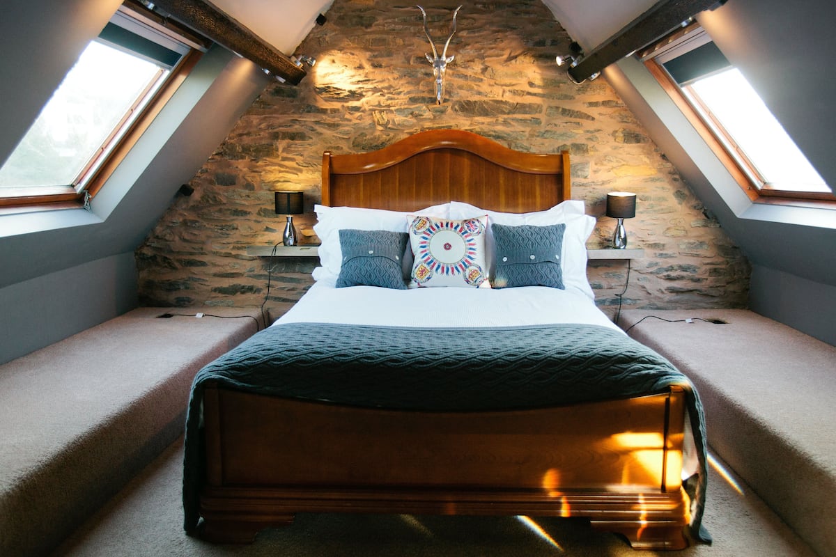 A bedroom with a sloped ceiling, stone wall, wooden bed frame, and two bedside lamps.