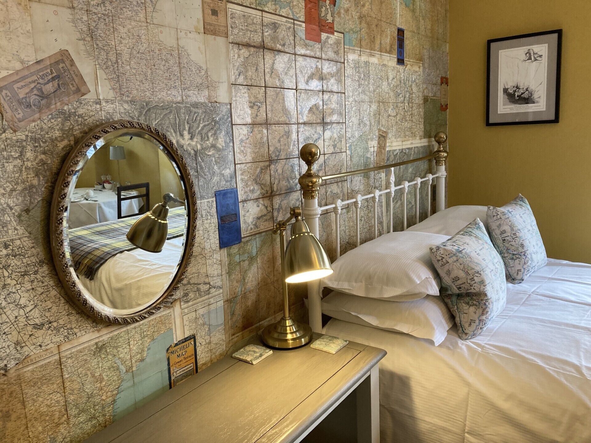 A neatly made bed with white linens and patterned pillows, a round mirror with a decorative frame, a brass desk lamp, and a map-adorned wall.