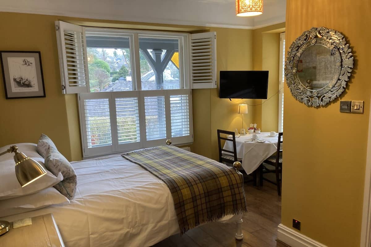 A bedroom with a bed, a TV mounted on the wall, a dining table with chairs, and a window with shutters.