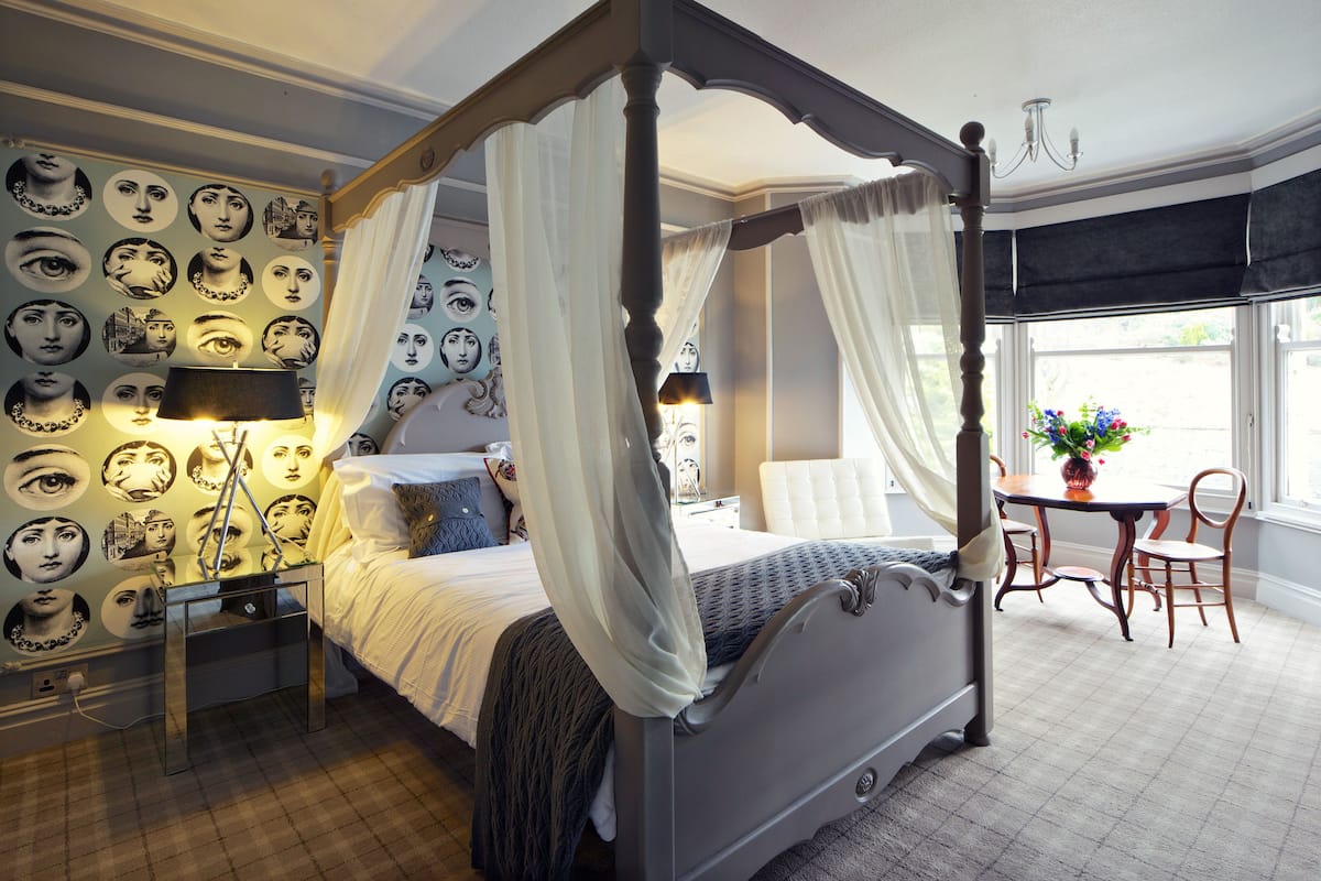 A four-poster canopy bed with a nightstand and lamp, a chair, and a table with flowers.