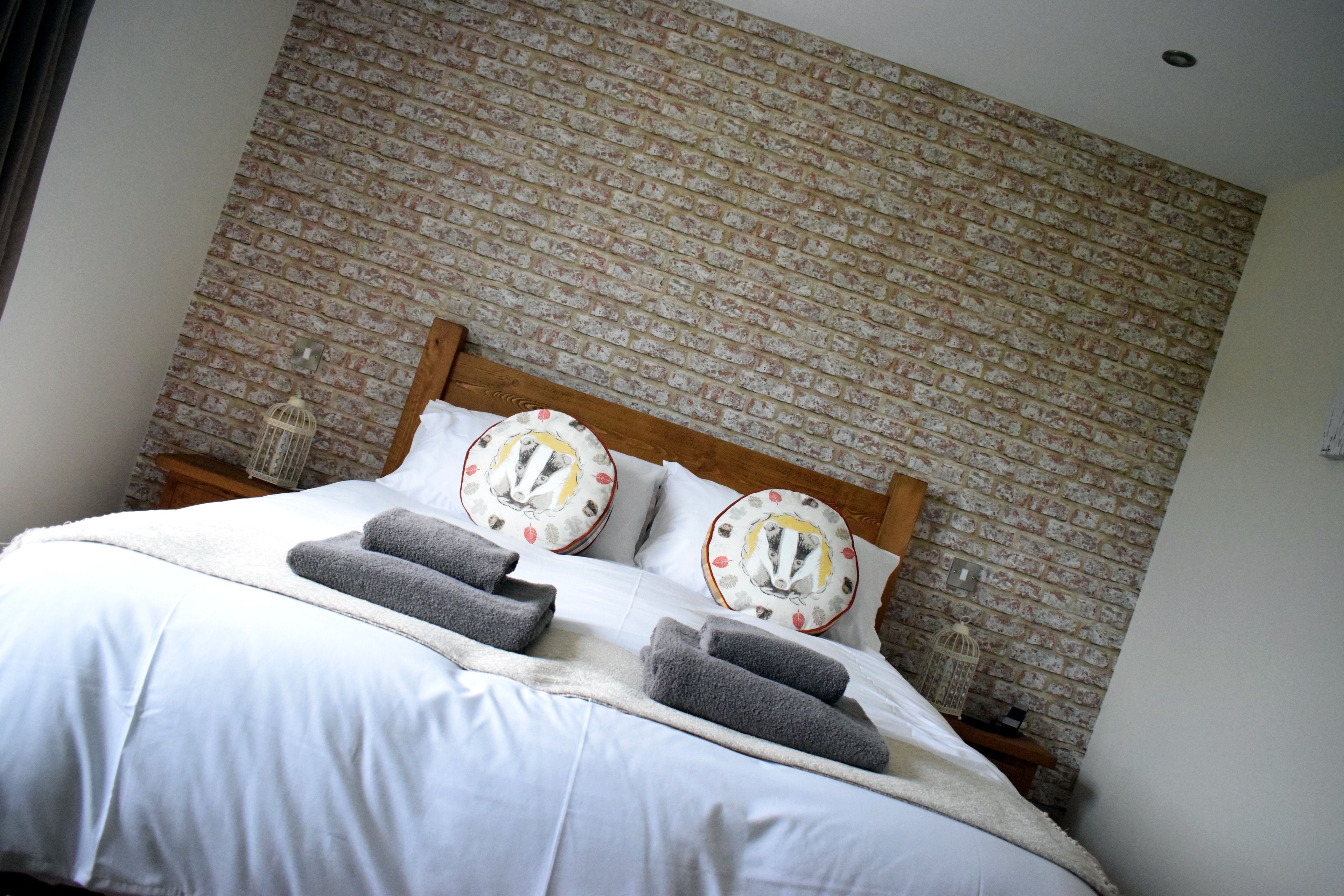 Una cama bien tendida con sábanas blancas y toallas grises, un cabecero de madera y una pared de ladrillo con diseño.