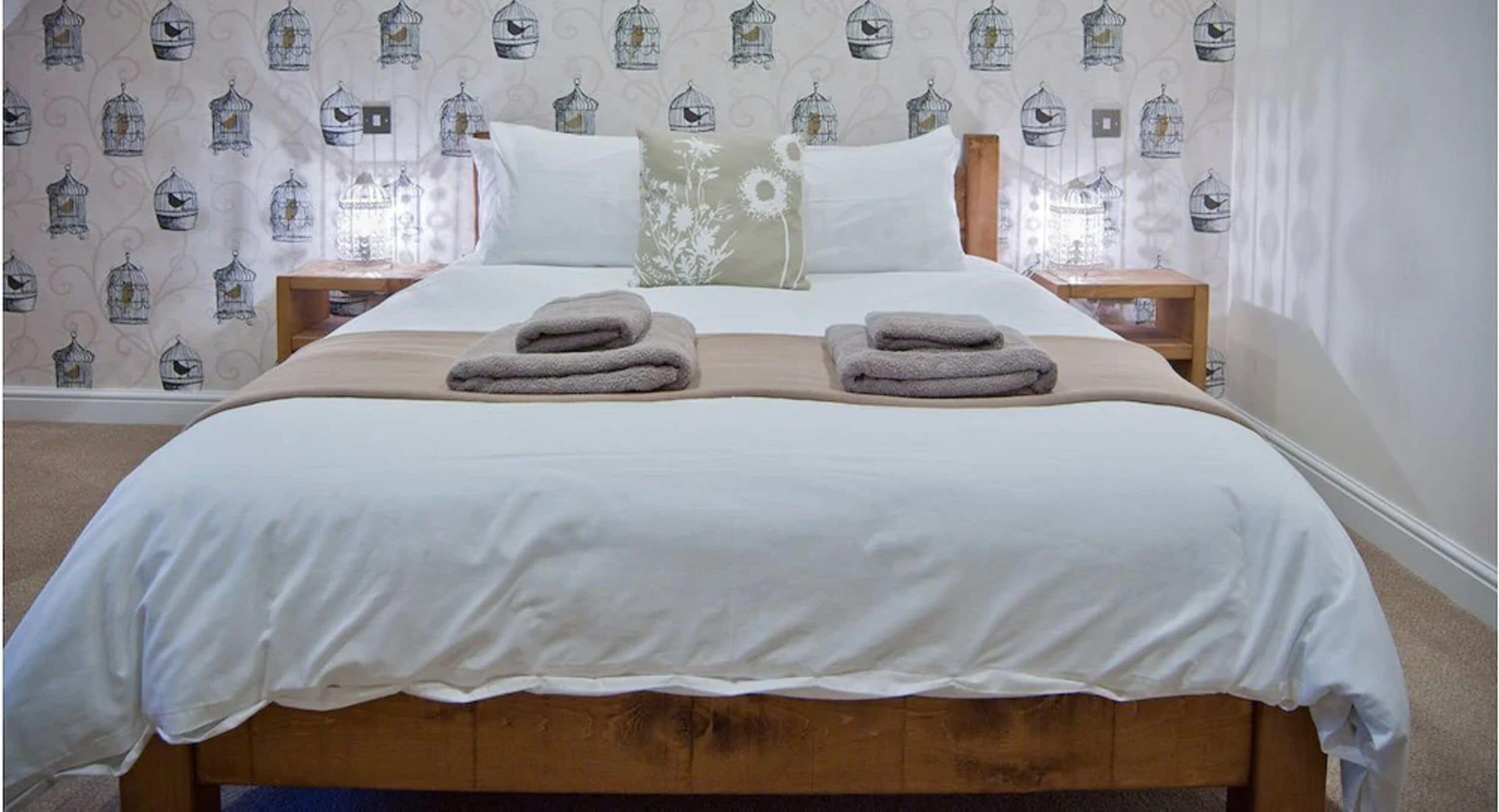 Una cama bien hecha con dos toallas dobladas, cabecera de madera y papel tapiz estampado con jaulas de pájaros.