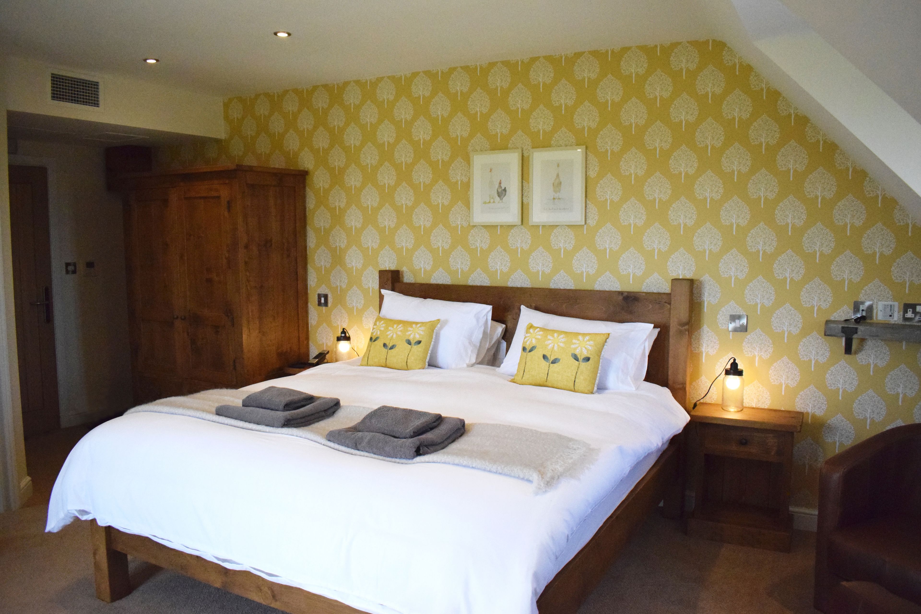 A neatly made bed with white linens and two pillows, a wooden wardrobe, and a nightstand with a lamp.