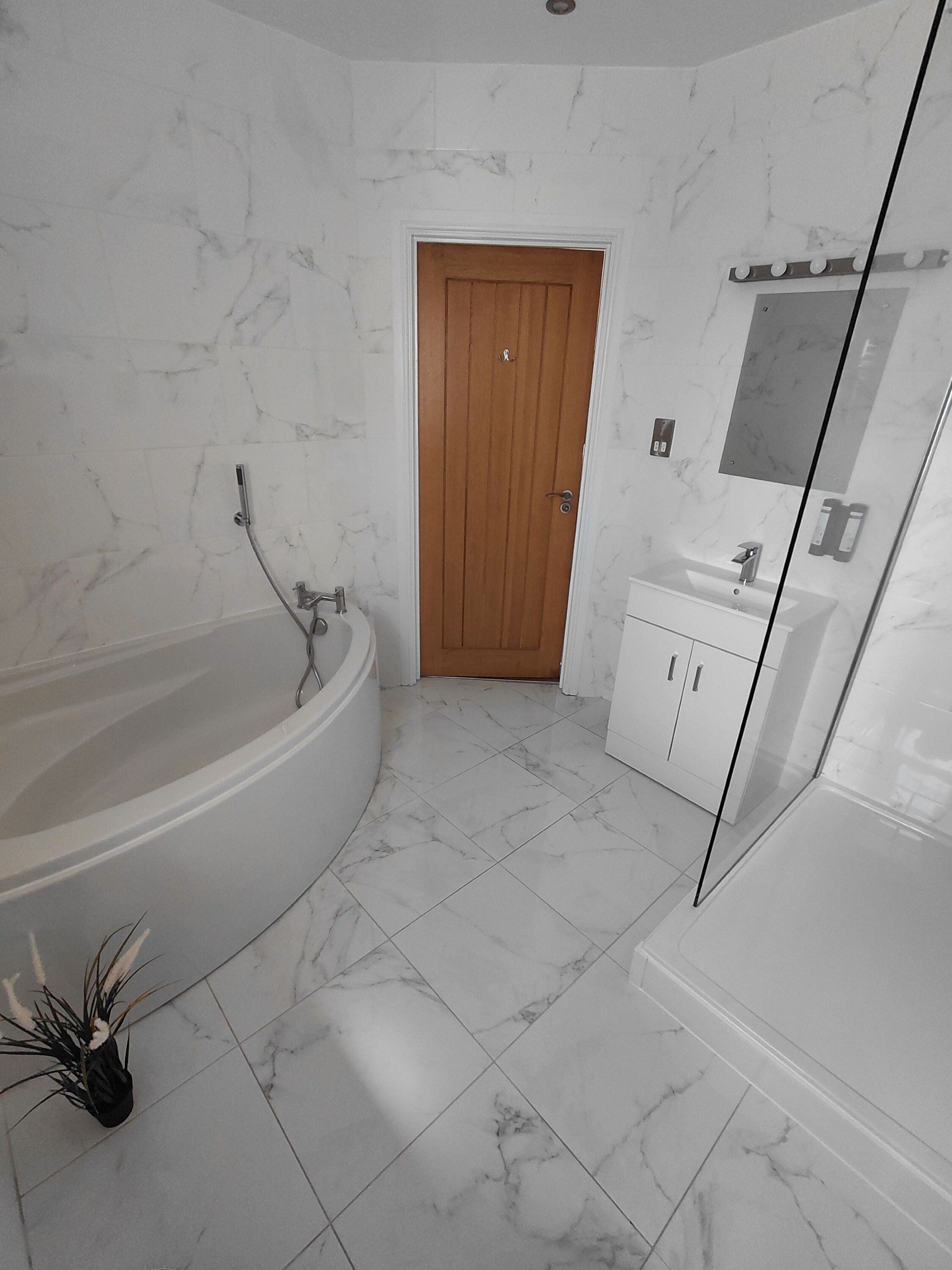 Un bagno moderno con una vasca freestanding, una porta in legno, un box doccia in vetro e un mobile lavabo bianco con specchio.