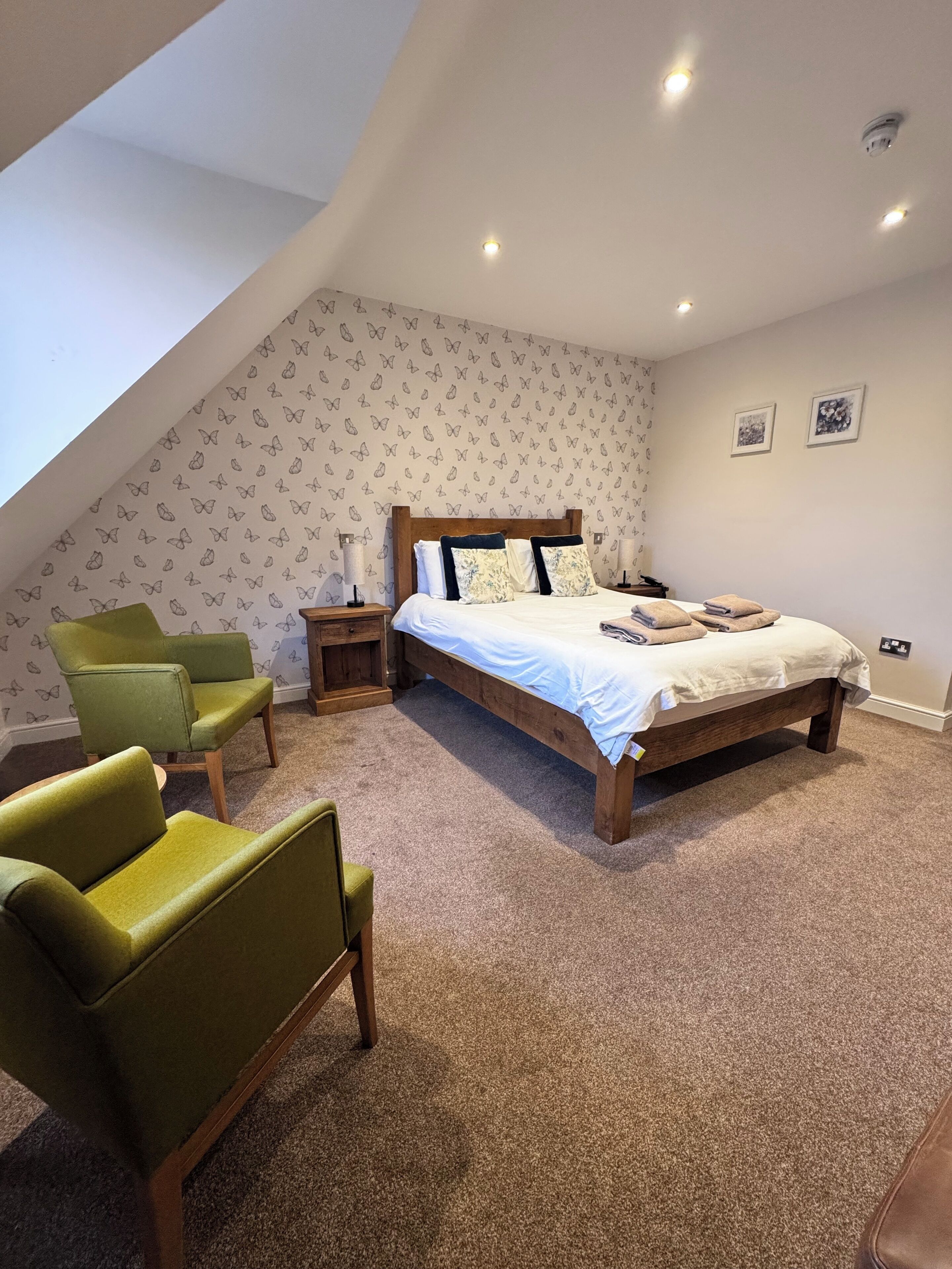 A bedroom with a sloped ceiling, a bed with a wooden headboard, two green armchairs, a nightstand, and a small table.