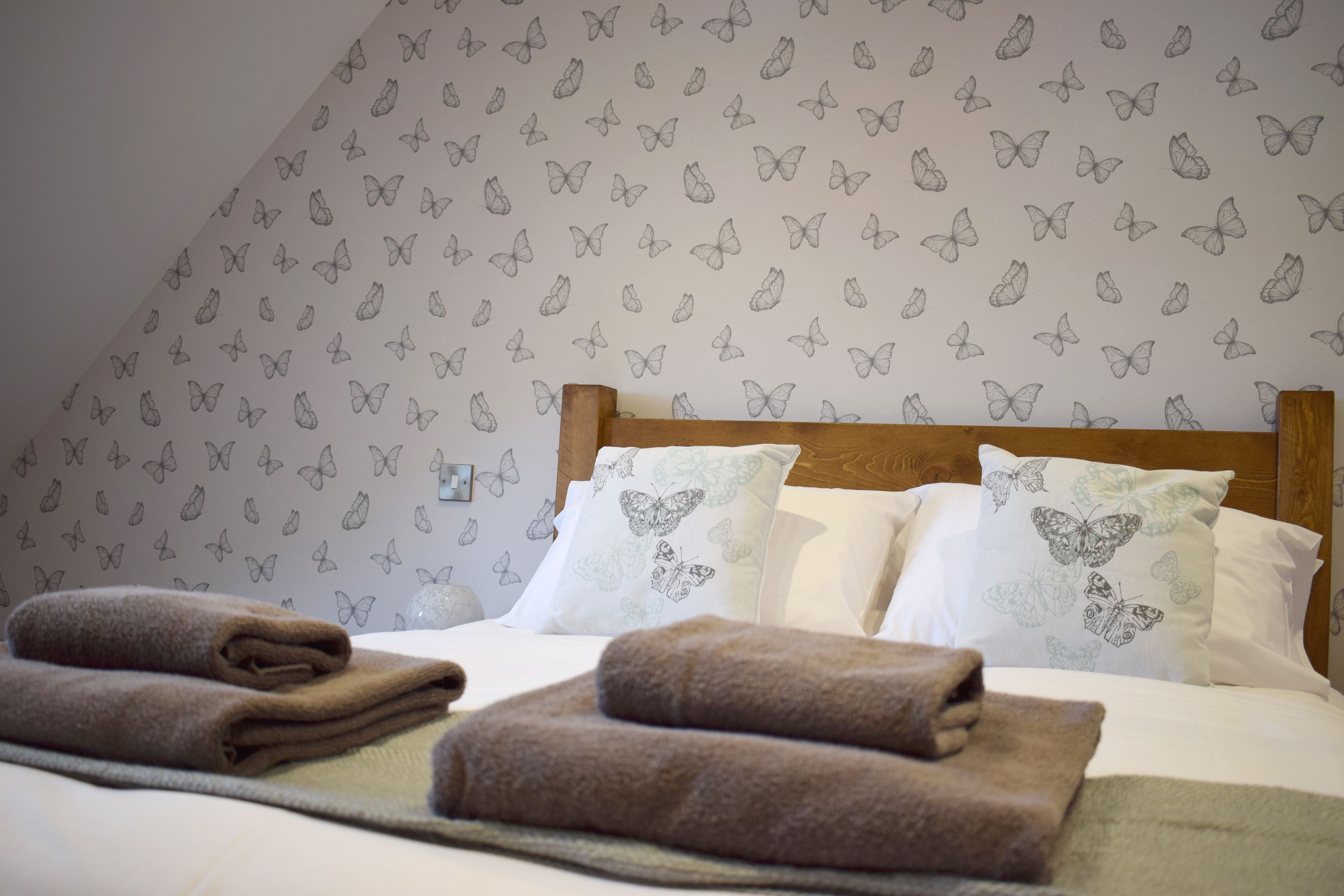 A neatly made bed with folded towels, a wooden headboard, and butterfly-patterned wallpaper.